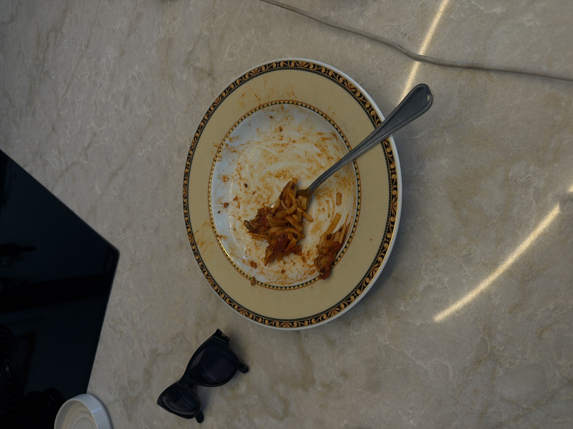 A close-up, high-angle shot shows a nearly empty plate of pasta with red sauce and a fork resting on a marble-patterned countertop. To the left of the plate, a pair of black sunglasses are laid down. In the background, a black glass stovetop is visible, with a white circular ceramic dish to its left. The scene is located in Ridderkerk, Netherlands.