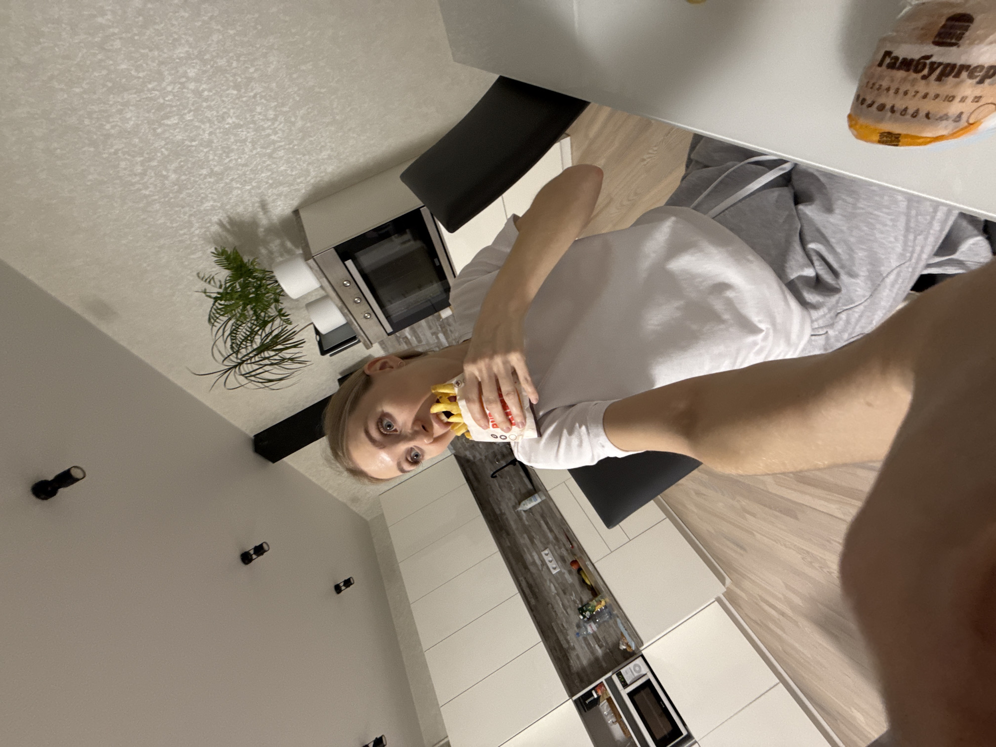A woman in a white t-shirt and grey sweatpants is eating french fries in a kitchen. A package labeled "ГаМбуРГеР" is on the table. The kitchen has white cabinets, a black countertop, and a black chair. A microwave is built into the lower cabinets. A plant is visible atop an upper cabinet. Black track lighting is on the white wall. Based on the Cyrillic script and the architectural style, the location is likely Izhevsk, Russia.