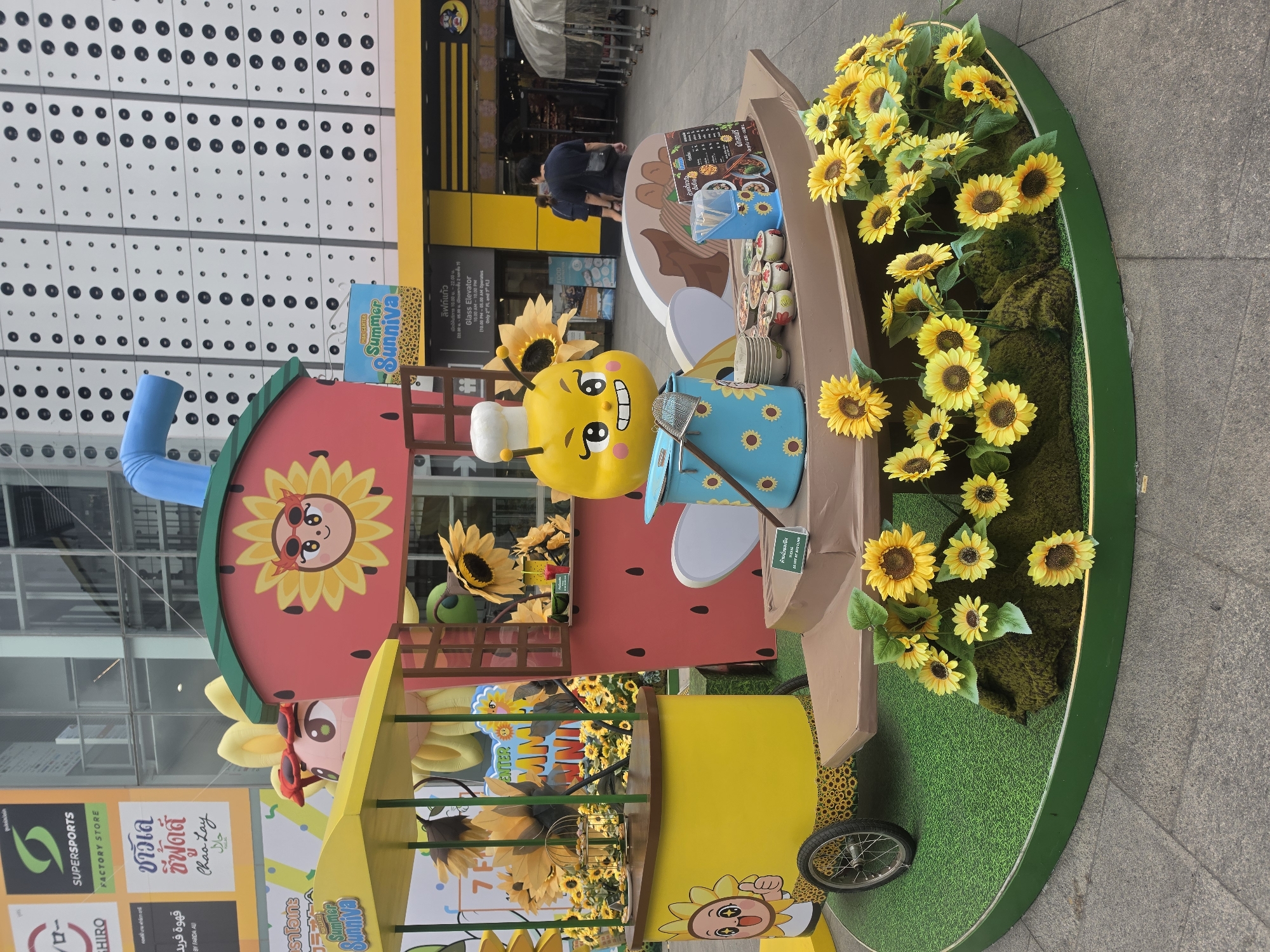 The image depicts a colorful and elaborate sunflower-themed installation likely used for promotional purposes, positioned on a sidewalk in an urban environment. The installation features cartoon-like characters, including a bee wearing a chef's hat, sunflowers in pots and planters, and structures resembling food carts or stalls. The structures are painted in bright colors, with details like sunflower faces and watermelon designs. The surrounding environment includes a building with a white facade dotted with black circles and another building with yellow signage. A man wearing a dark blue shirt is walking on the sidewalk. The location is Bangkok, Thailand, as evidenced by the Thai script on the store signs visible in the background. Specifically, signs for "Supersports Factory Store", "Chao Lay Seafoods", and "Summer Sunniva" are present.