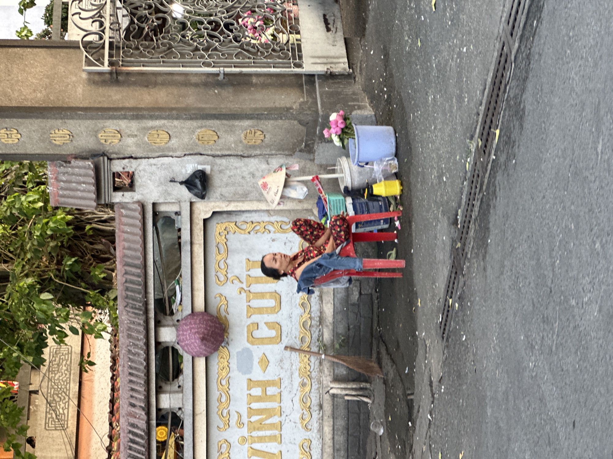 A woman sits on a red plastic chair on a sidewalk in Thủ Đức, Vietnam. She is wearing a floral print top and blue pants. Her head is resting against a wall, suggesting she is resting or sleeping. Next to her are various containers, including a blue bucket filled with pink and white flowers, and other assorted items. Behind her is a building with ornate gold lettering that appears to read "Vinh Cú". A broom leans against the building nearby. The sidewalk is wet and leads to a street.
