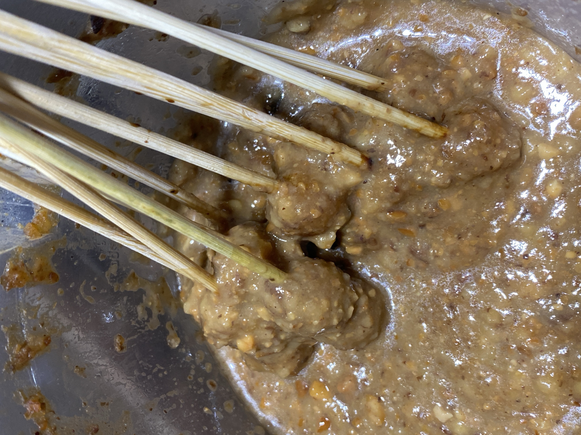 A close-up shot captures pieces of food skewered on bamboo sticks, submerged in a creamy, peanut-based sauce. The food appears to be small, round meatballs or chunks of meat, coated in the sauce. The sauce itself is a light brown color and has a slightly oily sheen. Some small, crushed peanut pieces are visible within the sauce. The background is a metallic pot or bowl, with some remnants of the sauce and food clinging to its sides. The lighting suggests an indoor setting, likely a kitchen. This scene could be found in a food stall or home kitchen in the city of Kasihan, Indonesia, where dishes like satay are commonly prepared.