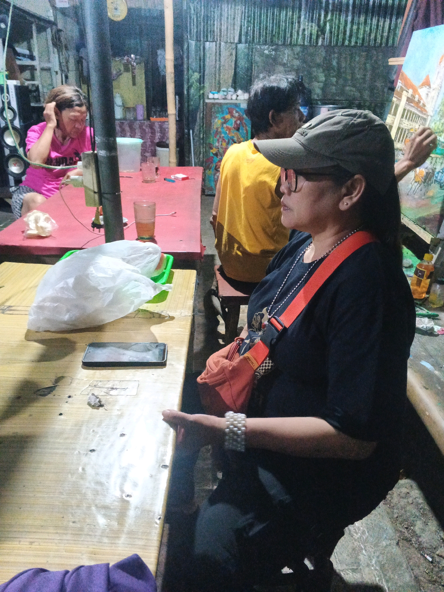 The image depicts an indoor scene, possibly a workshop or informal gathering space, with three individuals visible. In the foreground, a woman wearing a black t-shirt, an orange crossbody bag, and a green baseball cap is seated at a wooden table, looking to her right. She has a pearl bracelet on her left wrist. To her left, a white plastic bag containing items is placed on a green tray on the same table. A smartphone is also visible on the table. In the background, to the left, another woman in a pink t-shirt is seated at a red table with drinks and a lighter. Further back, a person in a yellow t-shirt stands near an easel with a painting. The environment appears cluttered, with various art supplies and building materials visible, suggesting an informal workspace. The lighting is dim and artificial. The scene is set in Ungaran, Indonesia, implying a local context for the depicted activities.