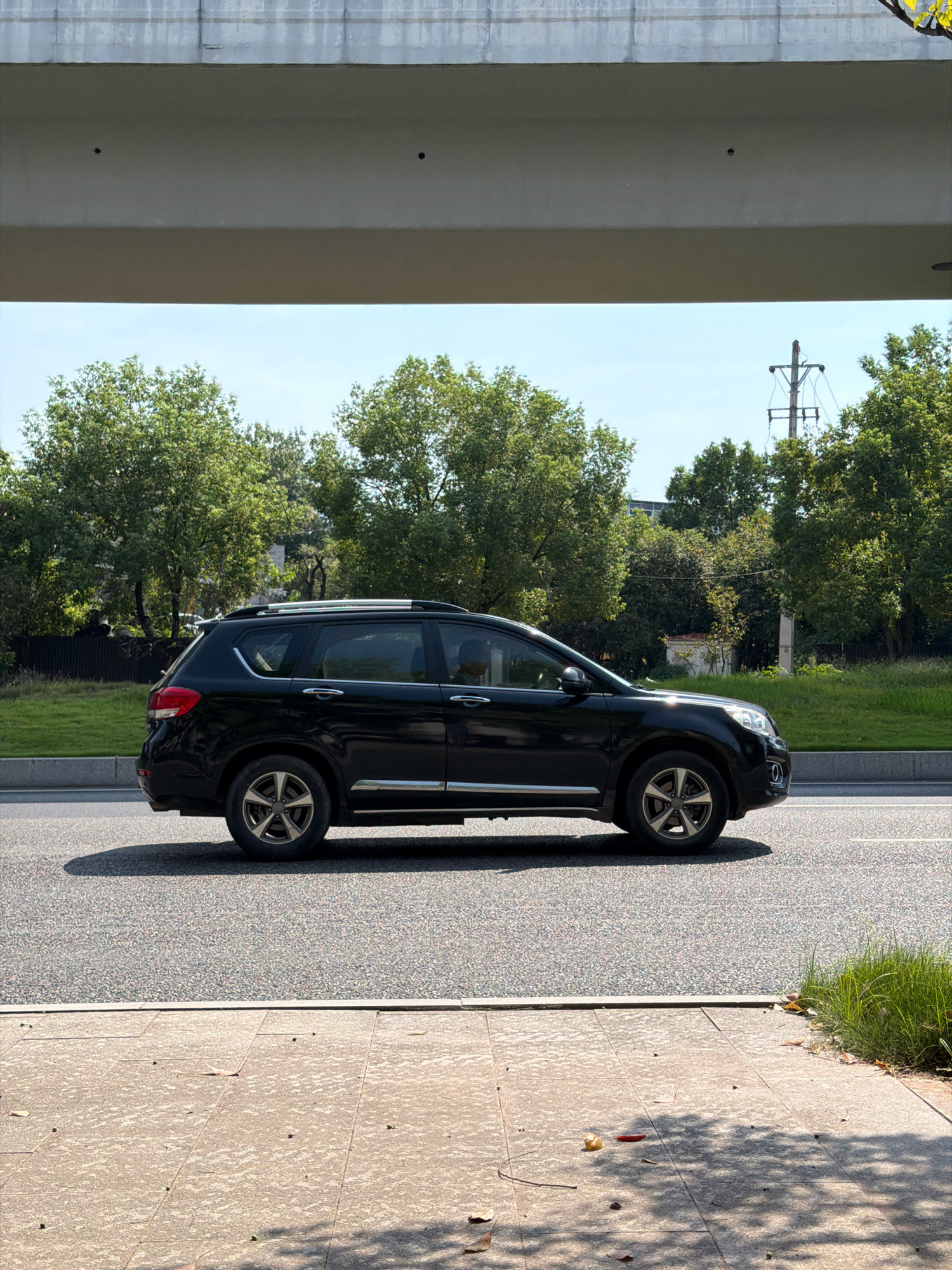 A black SUV is driving on a road under a concrete overpass in Jiangjin, China. The SUV is in the right lane of a two-lane road, with its front facing to the right. The SUV has a silver roof rack and silver trim around the windows. The road is asphalt and has white lane markings. To the left of the road is a sidewalk made of concrete pavers. Behind the road is a grassy embankment with trees. In the background, there is a utility pole with wires and a blue sky with scattered clouds.