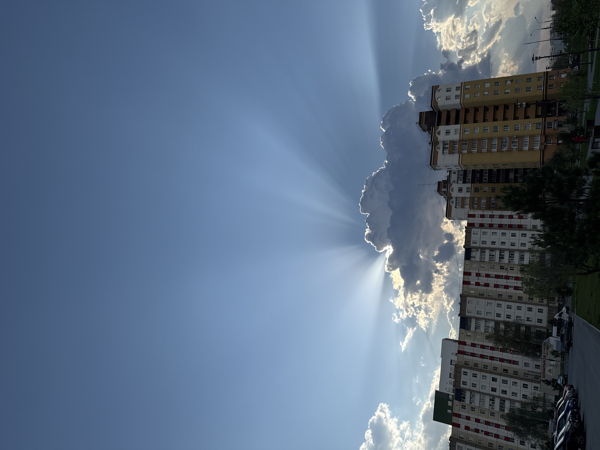 The image shows an urban scene in Tobolsk, Russia, featuring multi-story residential buildings adjacent to dramatic cloud formations. Sunlight breaks through the clouds in striking rays, casting light and shadow across the scene. The buildings vary in color, with some sections appearing yellow and others white with red accents. The sky is predominantly blue, contrasting with the bright white and gray of the clouds. A street with parked cars and greenery is visible along the bottom of the frame. The image is taken from a low angle.