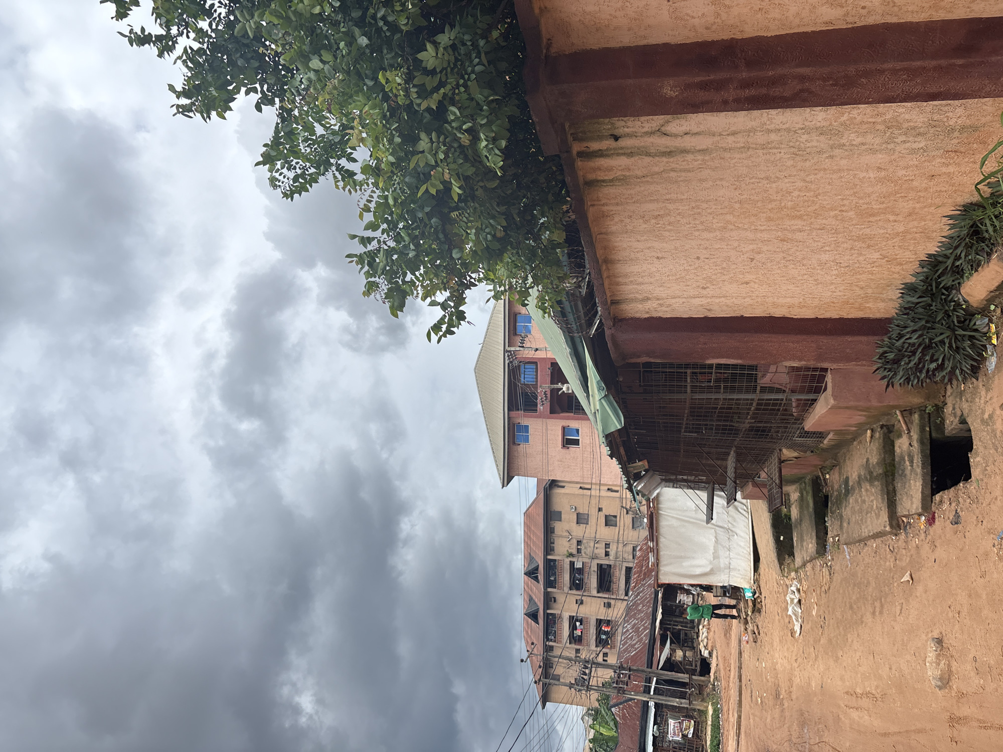 The image depicts a street scene in Nkpor, Nigeria, under a dramatic, overcast sky filled with dark, heavy clouds. In the foreground, a rough, sandy dirt road leads into the scene, with evidence of debris and a broken concrete drain cover. To the right, a textured, peach-colored wall with darker brown vertical accents dominates the frame, with a large green tree growing over its top. Behind the wall, a caged structure with metal shelves is visible, along with a green awning.
Further into the scene, on the left side of the road, is a white tent-like structure and various market stalls with corrugated metal roofs. A person in a bright green shirt and dark pants stands near the white tent, facing away from the camera. Behind the stalls, several multi-story buildings are visible, one a tan building with a red tiled roof and many windows, and another pink building with blue-framed windows. Electrical wires crisscross the sky above the buildings. The overall impression is of an urban environment in a tropical climate, possibly experiencing or anticipating rainfall due to the sky conditions.