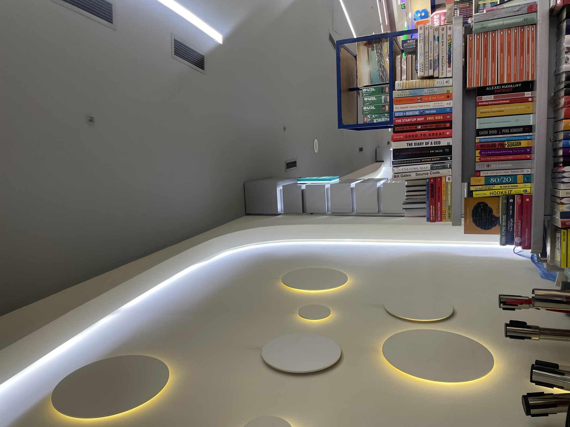 This media file captures a modern, bright interior space, likely a bookstore or a contemporary library, taken from a low angle. The scene is dominated by a clean, white aesthetic. A wide, curved white surface, possibly a counter or a platform, extends across the lower and mid-ground. This surface features multiple circular cutouts, each subtly backlit with a warm, yellow glow, creating a striking visual pattern.
Above this counter, linear LED strip lighting illuminates the space from the ceiling or upper wall, providing cool white light. To the right, several bookshelves are packed with books, arranged both horizontally and vertically. One section of books is contained within a blue-framed shelf unit. The upper wall is plain white, interrupted by two rectangular ventilation grilles and a small, circular device, possibly a smoke detector or sensor. No people are visible, and the setting appears quiet and well-maintained.
Visible text on the book spines includes titles such as "OPTION B," "HOOKED," "the everything store brad stone," "ORIGINALS," "THE 5 AM CLUB," "The Wealth Money Can't Buy," "KOTLER MARKETING," "THE STARTUP WAY ERIC RIES," "CLOCKWORK ORANGE," "GOOD TO GREAT," "THE DIARY OF A CEO," "THE CHANGING WORLD ORDER RAY DALIO," "BEYOND ORDER," "The CULTURE MAP," "Source Code," "SCRUM," "ZERO ONE," "LEADERS EAT LAST," "ALEXEI NAVALNY PATRIOT," "Branding that means Business," "POLITICAL ORDER AND POLITICAL DECAY," "SHOE DOG PHIL KNIGHT," "PRE-SUASION," "How to Stop Worrying and Start Living," "21 Lessons," "think and grow rich Napoleon Hill," "80/20," and "HOOKED." Authors like GRANT, LEONARDI, ROBIN SHARMA, ERIC RIES, ANTHONY BURGENS, JIM COLLINS, RAY DALIO, JORDAN B. PETERSON, Bill Gates, PETER THIEL, SIMON SINEK, FRANCIS FUKUYAMA, ROBERT CIALDINI, and NIR EYAL are also identifiable. Some book titles and other texts appear to be in Armenian, including "USUMԵԼ ԻՆՉՊԵՍ" and "ԴԵՆԻԵԼ ԳՈՒՄԱ," confirming the location context of Yerevan, Armenia. The books predominantly cover business, self-help, and non-fiction genres.