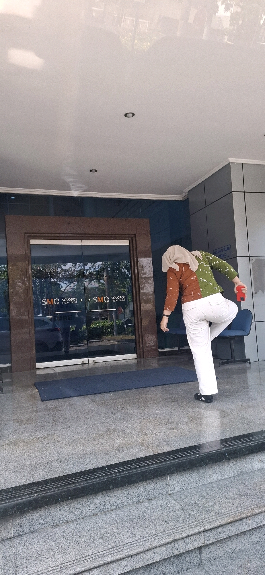 A person wearing a hijab, a long-sleeved multi-colored top, and white pants stands in front of a glass-paned building entrance. The person is holding a red object, possibly a phone, in their right hand and appears to be in the middle of a stretching or yoga pose, with their left leg bent and raised. The entrance doors are double glass doors with brown frames, and the building's logo, "SMG SOLOPOS MEDIA GROUP," is visible on the glass. A dark blue mat is placed on the granite floor in front of the entrance. The background shows a reflection of trees and buildings in the glass doors. A blue chair is visible to the right of the person. The scene appears to be in a public area, possibly an office building entrance, in Surakarta, Indonesia, as indicated by the "SOLOPOS" name, which is a local newspaper in Surakarta.
