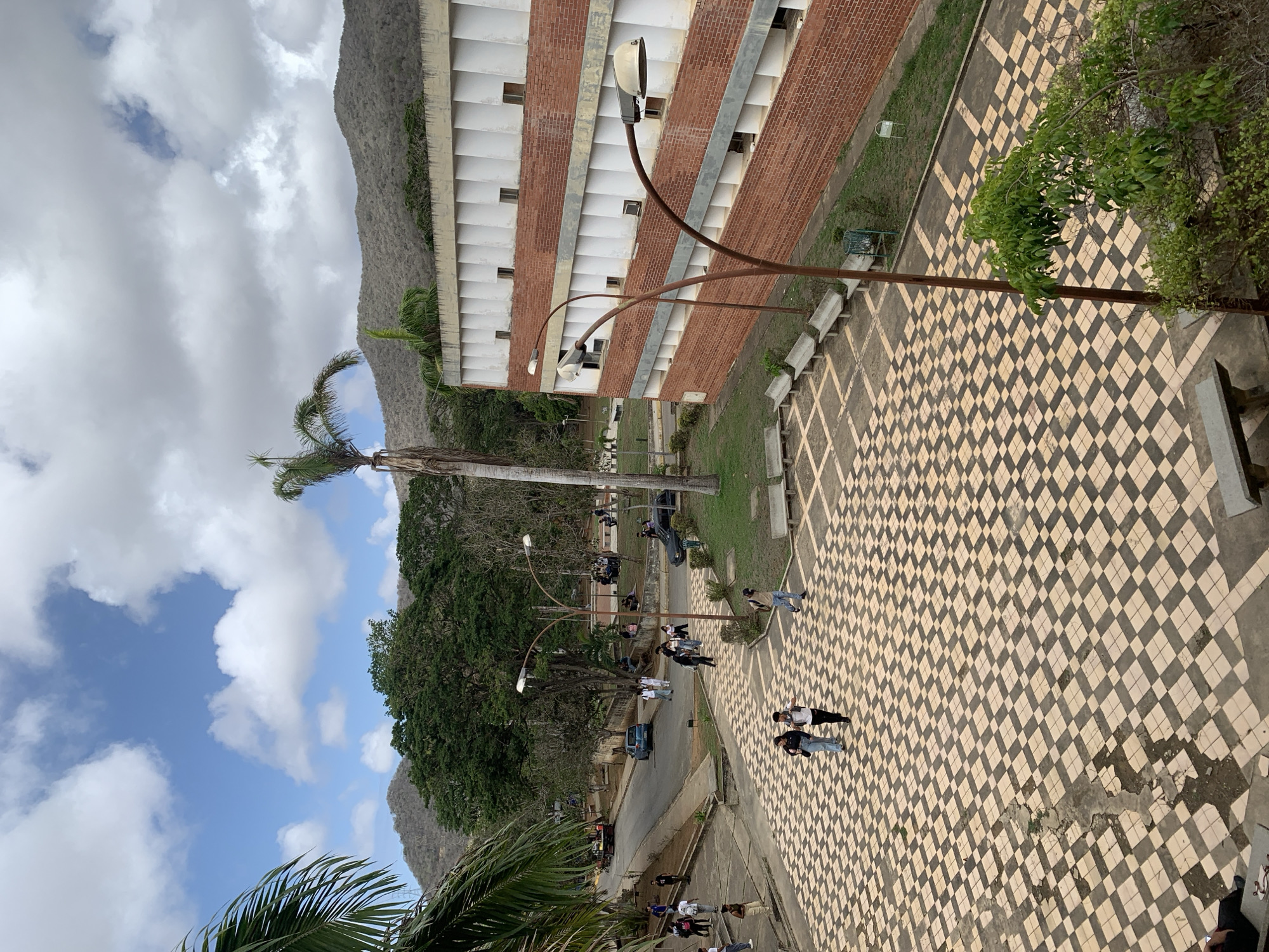 This image depicts an outdoor scene at a university campus, possibly located in Las Piedras, Venezuela. In the foreground, a large tiled courtyard with a checkered pattern of light beige and dark gray squares is visible. Several concrete benches are placed around the courtyard. Two individuals are walking across the tiled area. Further back, a paved pathway leads to a road where a blue car is parked. People are seen walking on the pathway and along the road. To the right of the courtyard, a long, multi-story building with a brick base and white upper sections with numerous vertical indentations is prominent. A tall palm tree stands in front of this building, and several street lamps with curved poles are also present. The background features lush green trees and dry, mountainous terrain under a partly cloudy sky. The overall impression is of a bustling academic environment on a sunny day.