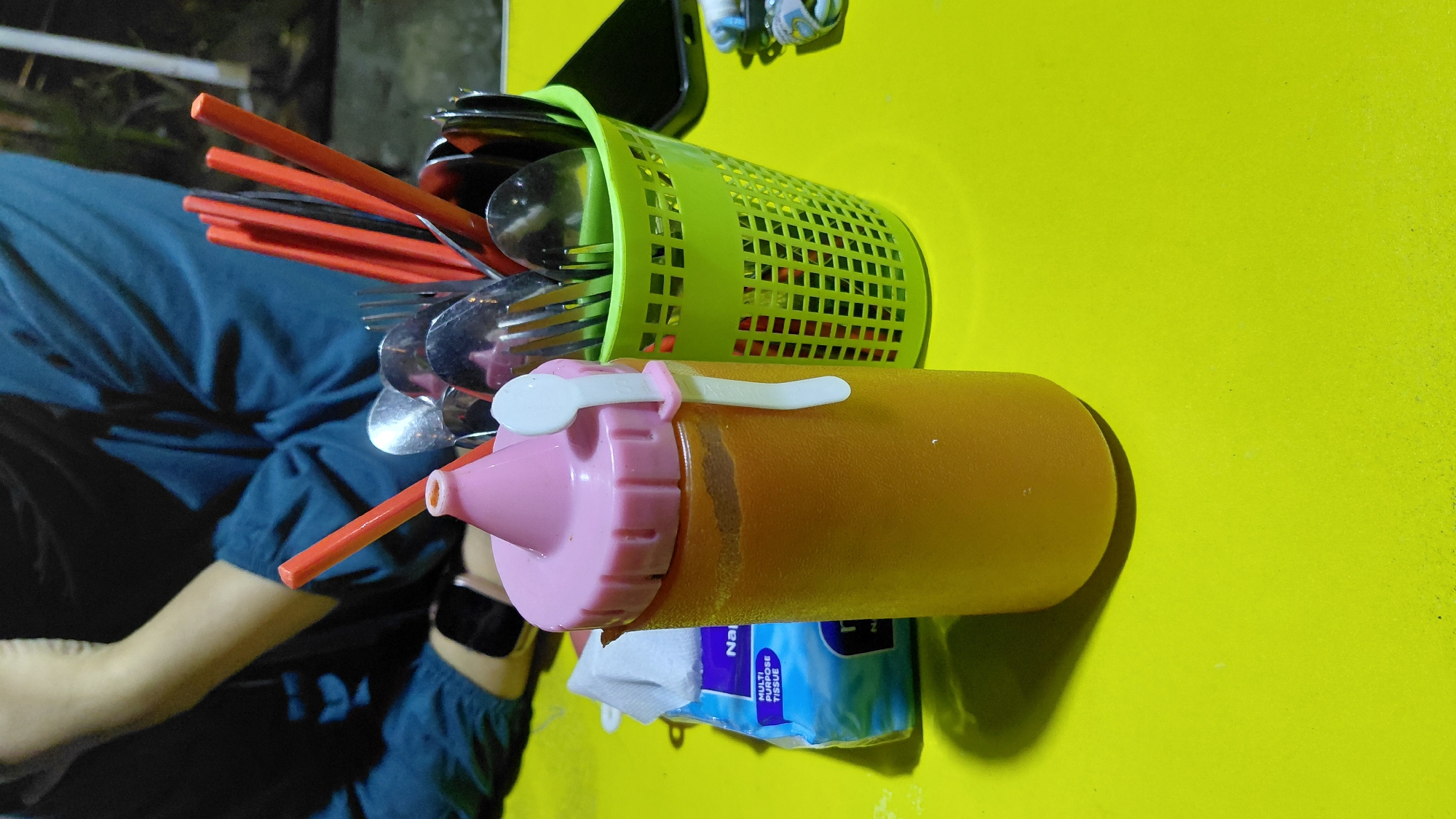 The image depicts a close-up view of a table set for a meal, likely in Bantarsoka, Indonesia. In the foreground, a squeeze bottle with a pink lid contains an orange liquid, possibly a sauce or condiment. A white plastic spoon is attached to the side of the bottle by a pink clip. To the right of the bottle, a bright green plastic basket holds an assortment of cutlery, including spoons, forks, and chopsticks. Some of the cutlery are red, likely chopsticks, sticking out of the basket. In the background, a person wearing a dark blue long-sleeved shirt is partially visible, with their arm resting on the table. A blue tissue packet is positioned to the left of the squeeze bottle. The table surface is a vibrant yellow. The lighting suggests an indoor or evening setting.