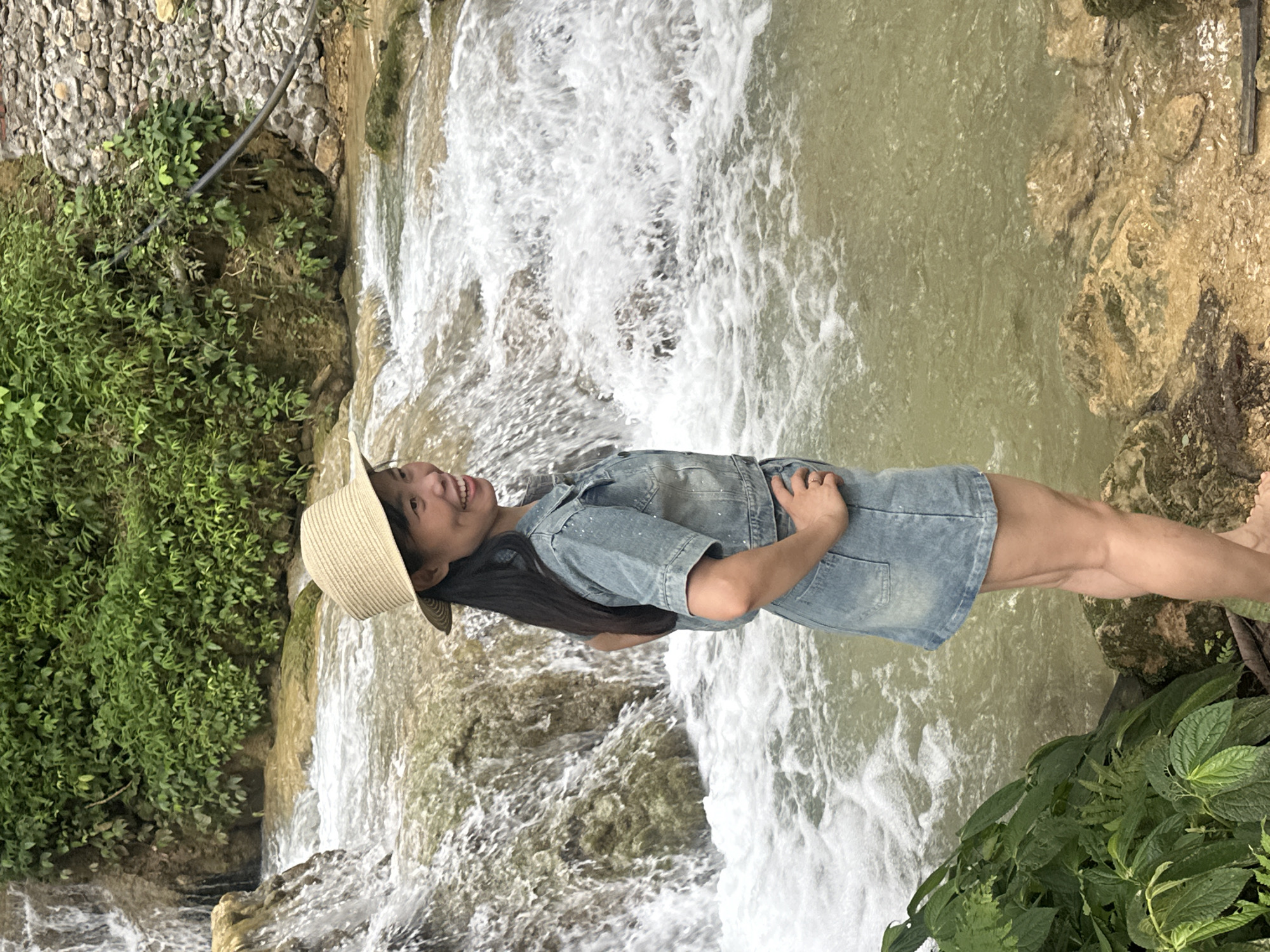 A medium shot captures a woman standing on rocks by a waterfall in Cổ Lũng Commune, Vietnam. She is wearing a straw hat, a denim jacket, and a denim skirt, and is smiling. The waterfall cascades down rocks in the background, with lush green foliage on either side. The water is clear and flowing, with white foam on the surface. The woman's hands are placed on her hips. The overall scene is serene and picturesque, with the natural beauty of the waterfall and surrounding vegetation being prominent.