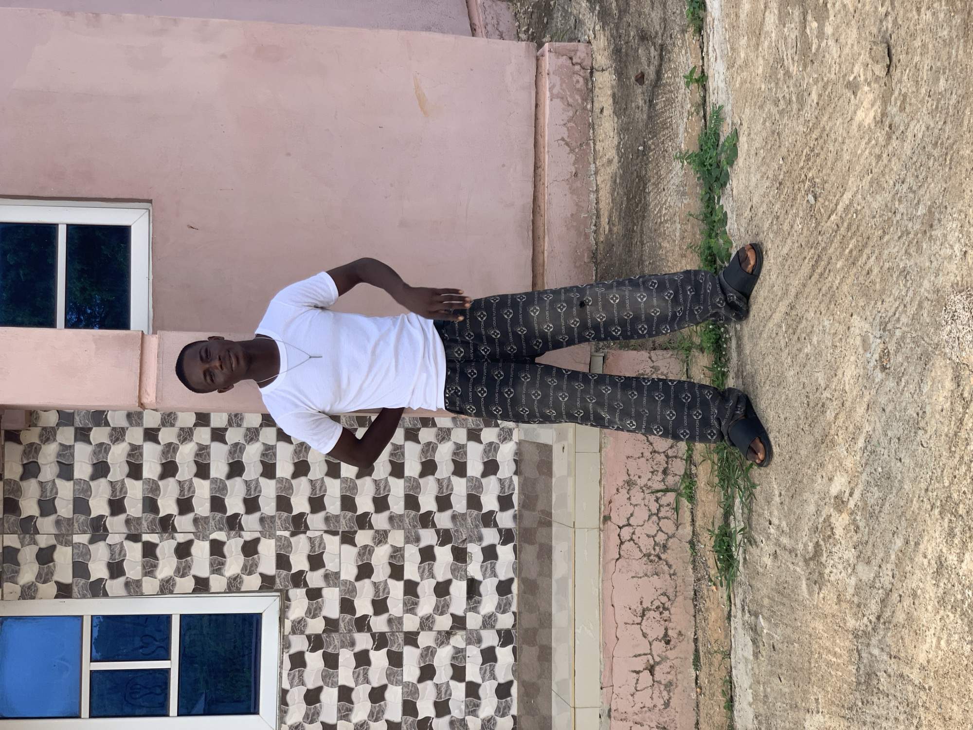 A young man stands in front of a building. He is wearing a white T-shirt, black pants with a repeated pattern, and black sandals. His hands are on his hips. Behind him is a tiled wall, a light pink wall, and a concrete walkway with patches of vegetation. A window is visible on the left side of the frame. The photo was taken in Gbeleko, Nigeria.