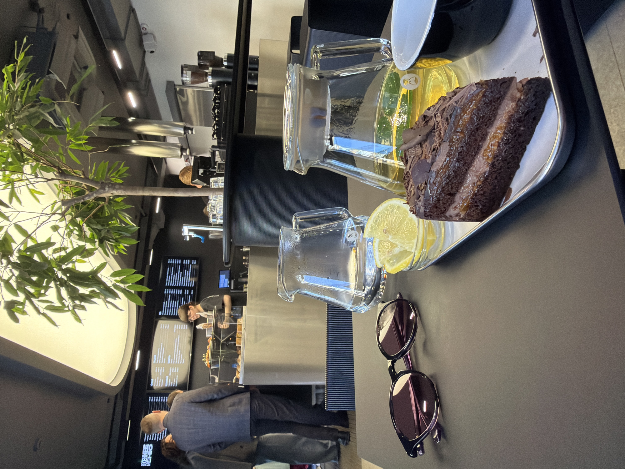 The image captures an indoor scene, likely a cafe or restaurant in Izhevsk, Russia. The focus is on a tray with a slice of chocolate cake, a glass carafe containing a yellow liquid (possibly a juice or lemonade) with a lemon slice, and a cup. Sunglasses are also placed next to the tray.
In the background, indistinct human figures can be observed, including an individual wearing a suit and possibly another person dressed in casual attire. A server is also visible behind a counter. The interior seems modern, with screens displaying menu items and greenery enhancing the ambiance.