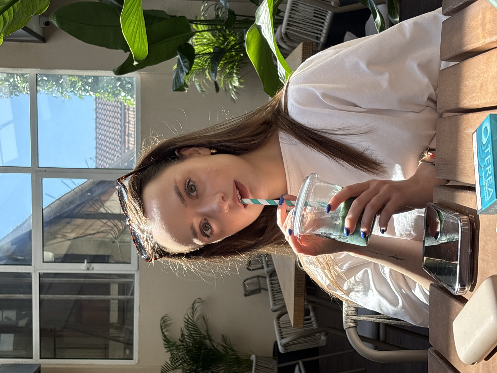 A young woman with long brown hair is seated at a wooden table outdoors, holding a clear plastic cup with a green beverage and a striped straw to her mouth. She is wearing a white t-shirt and sunglasses are pushed up on her head. Behind her, a window reveals a glimpse of buildings and a bright blue sky. Lush green plants are visible in the foreground and background. On the table in front of her, a smartphone and a small rectangular object are present. The setting appears to be a cafe or restaurant in Kragujevac, Serbia, given the presence of outdoor seating and an ambiance suggestive of a dining establishment in the city.