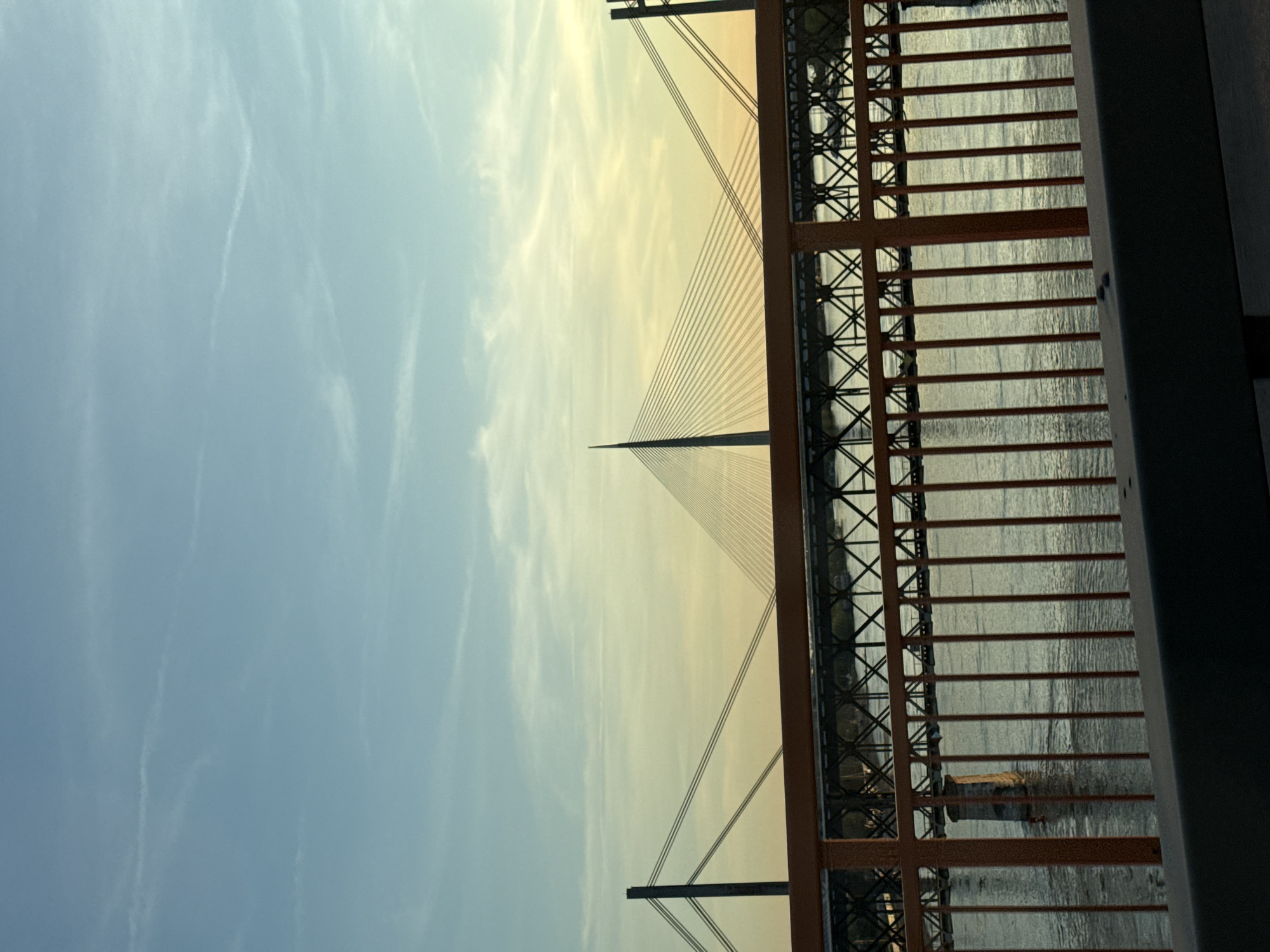 The image is an upward-angled view of the Ada Bridge in Belgrade, Serbia, as seen through an orange metal railing. The bridge's central pylon is visible against a light-blue and yellow sky with some wispy clouds. The bridge's suspension cables extend downwards from the pylon. Water, presumably the Sava River, is visible through the railing. The overall scene suggests a sunset or sunrise.