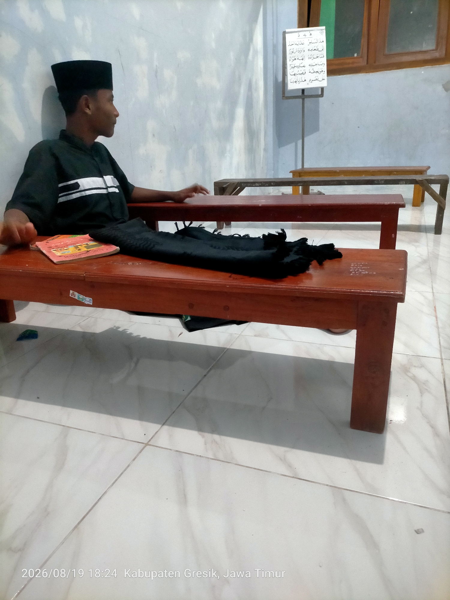 A young male wearing a black peci and a black shirt with white stripes is sitting on a wooden bench. He is looking to his right. On the bench next to him are a book and a black cloth. A whiteboard with Arabic text is visible in the background. The floor is tiled. The timestamp indicates the image was taken on 2026/08/19 at 18:24 in Kabupaten Gresik, Jawa Timur.