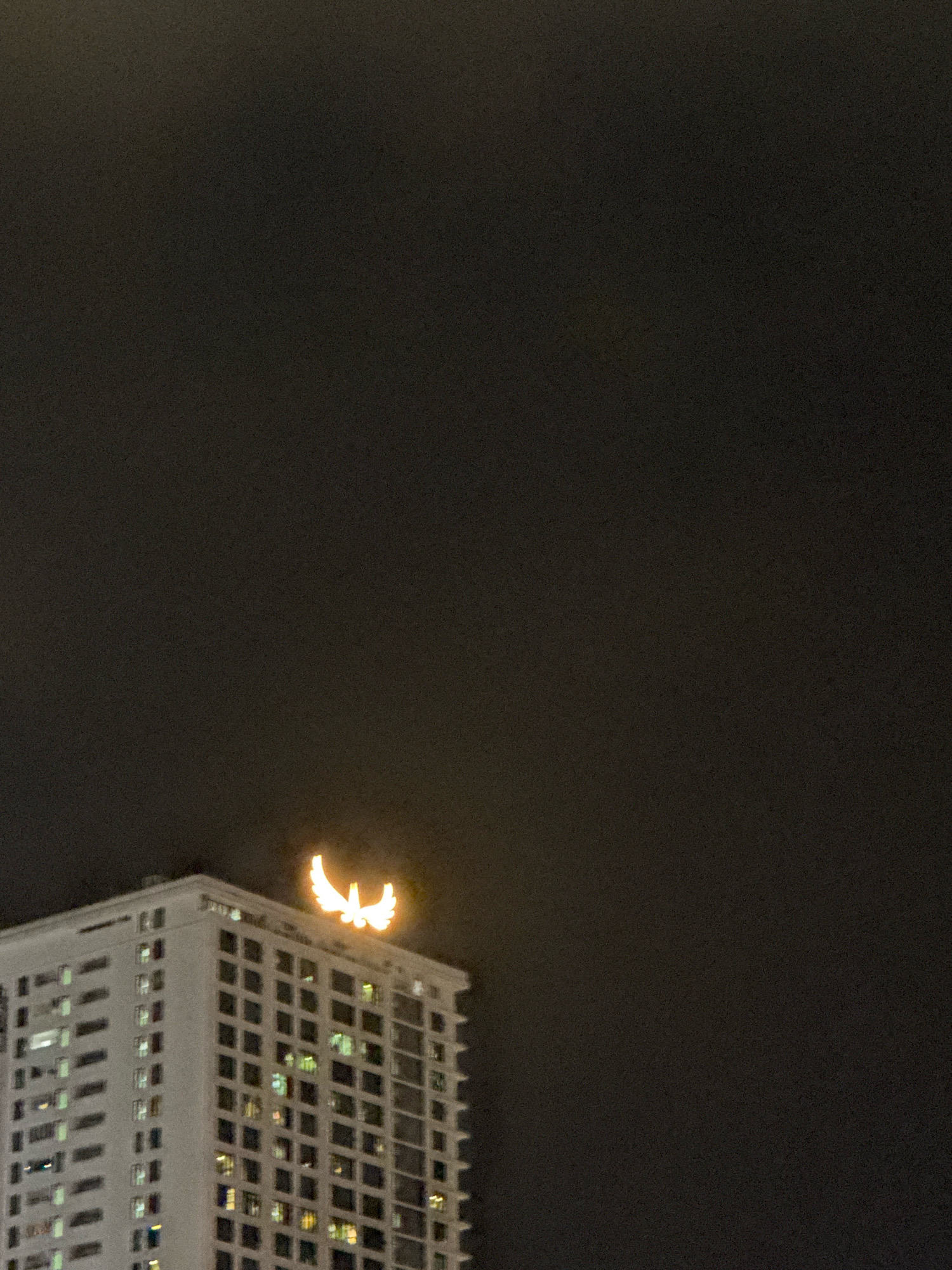 The image shows the top of a tall, white building at night. The building has many windows, some of which are illuminated, suggesting occupancy. At the very top of the building, there is a brightly lit, stylized logo that resembles a pair of wings or a phoenix. The background is a dark, featureless sky, indicating the image was taken after sunset or before sunrise. The overall scene is one of urban architecture illuminated against the night sky. The building appears to be the Muong Thanh Nha Trang Hotel in Nha Trang, Vietnam, based on the distinctive winged logo that is characteristic of the Muong Thanh hotel chain.