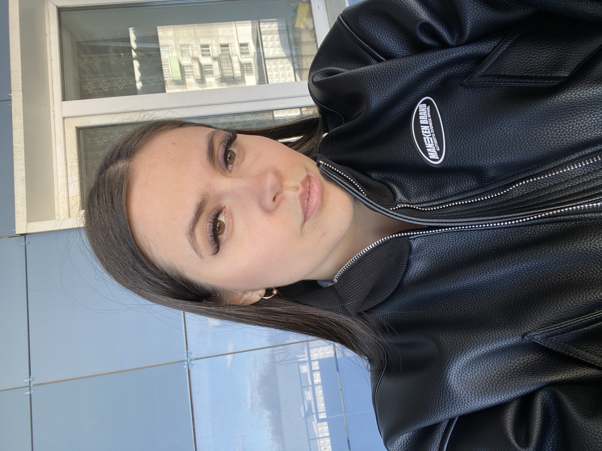 A young woman is posing for a selfie outdoors. She is wearing a black leather jacket with a zipper and a brand logo on the chest that reads "MANEKEN BRAND Authentic Oversize Apparel." She has long, dark hair and is wearing subtle makeup. She is facing the camera with a slight smile.
The background shows a building and blue tiled exterior. The location is in the city of Chaikovsky, Russia, indicated by the Cyrillic script on the windows visible in the reflection on the windows, and the architectural style. The environment suggests an urban setting. The image is slightly rotated, but the overall scene is well-lit.