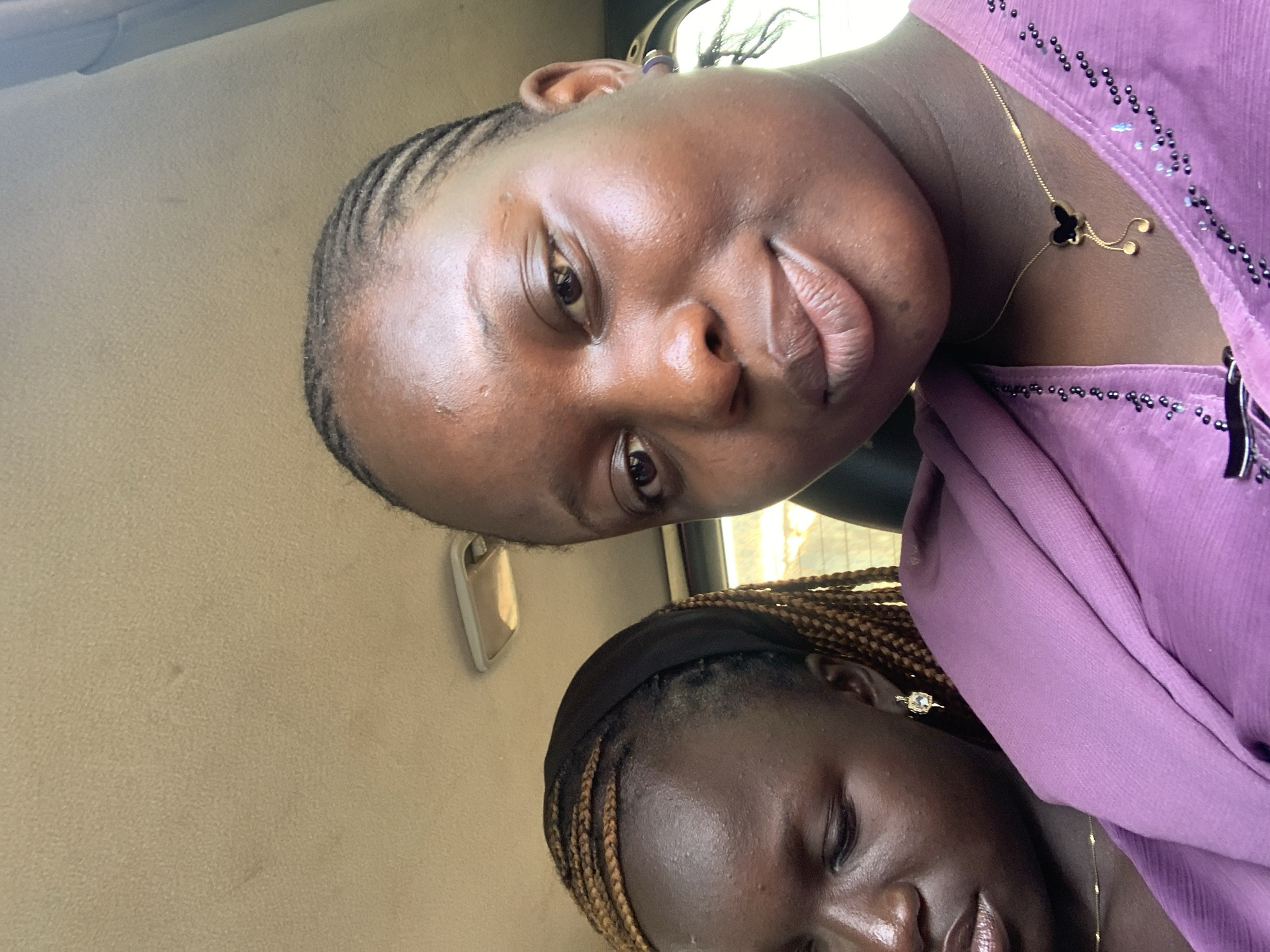 Two young Black women are seated in a car. The woman in the foreground is looking at the camera, smiling slightly. She has her hair braided in cornrows and is wearing a purple top with a butterfly necklace. The woman in the background is looking down and to her left. She has her hair braided into long braids and is wearing a black headband and a purple top. The car interior appears to be beige and the lighting suggests it is daytime. The exact location in the city of Unknown, Unknown cannot be determined from the image.