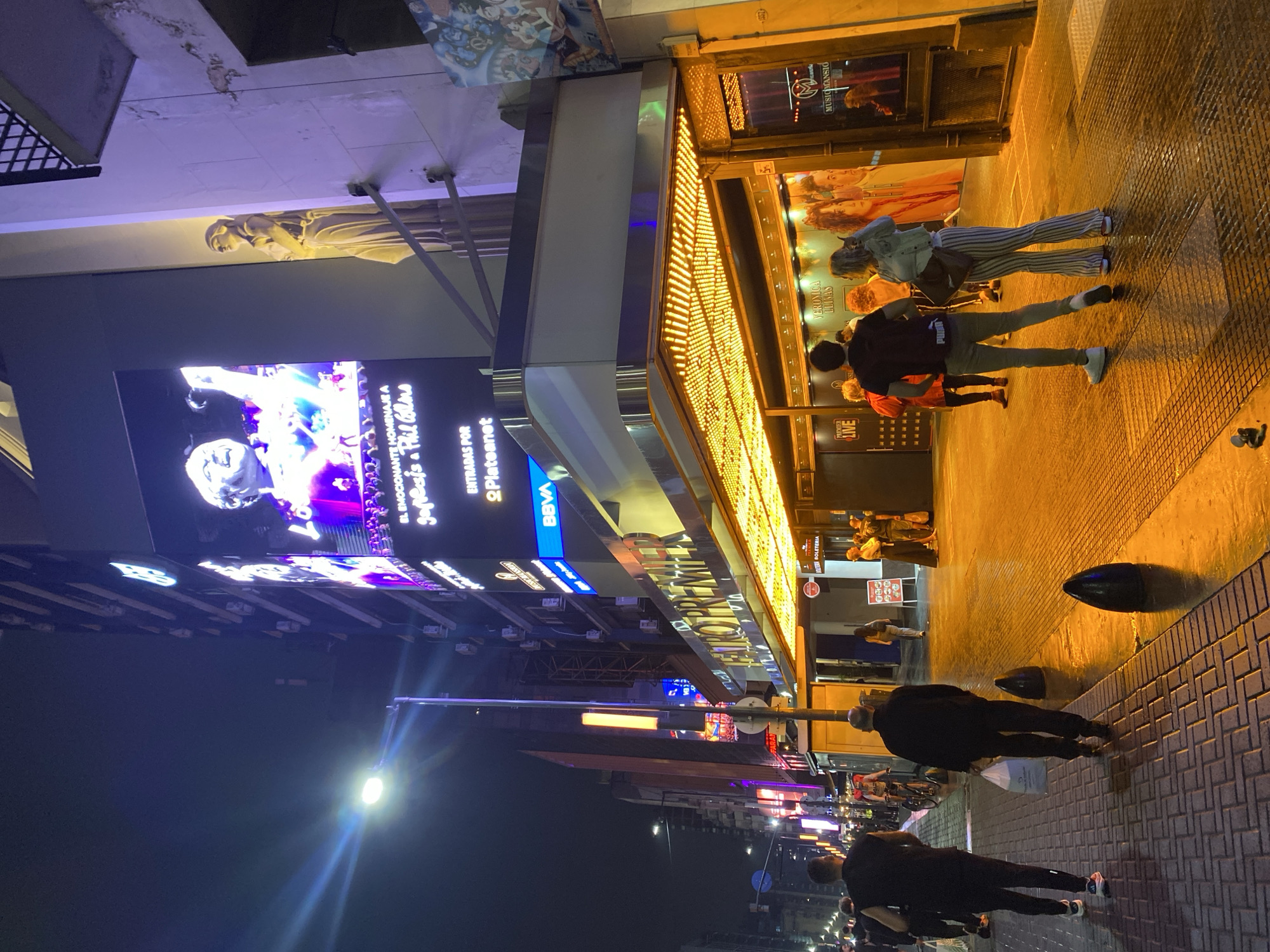 The image depicts a vibrant nighttime street scene on Avenida Corrientes, Buenos Aires, Argentina, centered on the Teatro Premier. The multi-story building features a large, vertically oriented digital billboard on its upper facade, displaying an illuminated image of a male performer and text reading "EL EMOCIONANTE HOMENAJE A Genesis y Phil Collins." To the right of the billboard, a classical-style female statue is affixed to the building's facade. Below the billboard, a prominent marquee with numerous small yellow lights extends over the theatre entrance, with "TEATRO PREMIER" illuminated above it. Other visible business signs include BBVA and Music Mansion, along with a "BOLETERIA" (ticket office) sign.
The street and sidewalk are illuminated by a combination of the theatre lights, the digital billboard's glow, and several bright streetlights, one of which is prominent on the left. The patterned brick sidewalk shows reflections from wet surfaces, as does the darker asphalt street. Two dark, conical bollards are present on the street's edge to the right.
Approximately fifteen individuals are visible. In the foreground on the left sidewalk, two men walk away from the camera; one wears a dark sleeveless shirt and dark pants, while the other, further ahead, wears a dark jacket and trousers, carrying a white plastic bag. Near the theatre entrance on the right, several people are gathered, including a man in a black t-shirt and grey trousers, and a woman in a light-colored jacket and striped bell-bottom pants. A cyclist is observed in the mid-ground on the street. The scene indicates active nocturnal urban pedestrian movement.