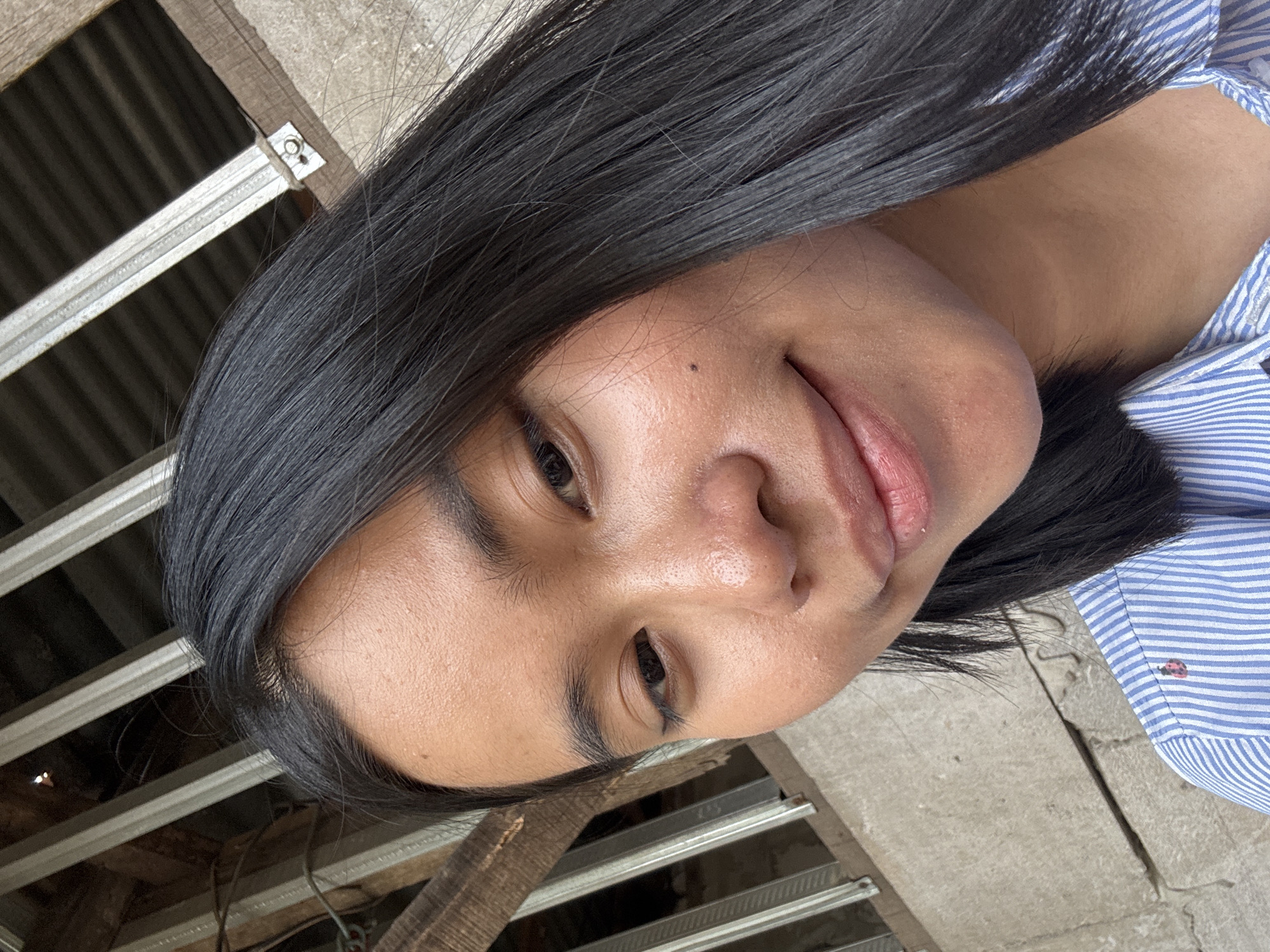 A young woman with long, dark hair is taking a selfie. She is wearing a blue and white striped shirt. The background appears to be outdoors with some wooden structures and a concrete wall. The lighting suggests it is daytime. There is a ladybug on her shirt. The location is identified as East Jakarta, Indonesia, though no specific landmarks or contextual clues in the image confirm this.