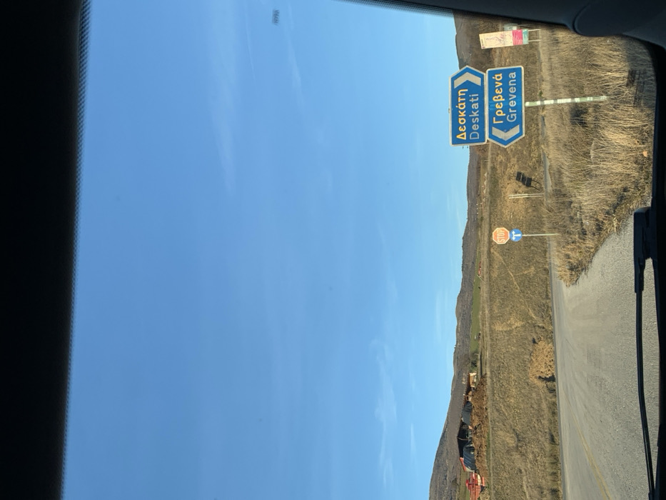 The image, taken from inside a vehicle looking through its windshield, captures a sunny daytime scene with a vast, clear blue sky overhead. The perspective is slightly rotated clockwise, showing the horizon line diagonally across the frame.
The central focus of the image is on two prominent blue directional road signs with white text, indicating destinations. The sign on the left displays "Δεσκάτη" (Deskati) in Greek and "Deskati" in English, suggesting a route forward or slightly to the right. The sign on the right, pointing more sharply to the right, reads "Γρεβενά" (Grevena) in Greek and "Grevena" in English. These signs are typical of road signage in Greece.
Below and to the right of these blue signs, a standard red and white international STOP sign is clearly visible. Adjacent to the stop sign is a circular blue mandatory instruction sign featuring a white arrow pointing to the left, indicating a required turn or direction.
The landscape beyond the road signs consists of dry, grassy hills and sparse vegetation, consistent with a rural or semi-arid environment. The road itself appears to be asphalt, curving off to the right. The bright, cloudless sky indicates clear weather conditions. The edges of the car's interior, likely the dashboard or windshield frame, are visible on the left and right sides of the image. The presence of the Greek town names "Deskati" and "Grevena" explicitly confirms the location as Greece, with the user context placing it around Grevena.