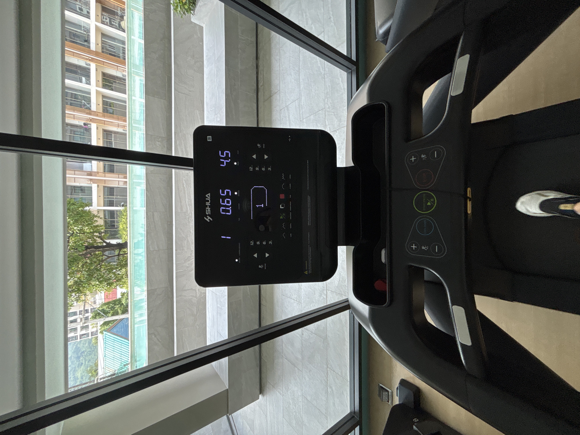 The image presents a vertical view inside a modern fitness area, likely a gym or a hotel fitness room, overlooking an outdoor scene. In the foreground, the console and part of the running belt of a black "SHUA" brand treadmill are visible. A person's right foot, wearing a white and black sneaker, is positioned on the treadmill belt, suggesting they are either actively running or about to start.
The treadmill's digital console is illuminated, displaying metrics such as "4.5" (likely speed), "0.65" (possibly distance covered, perhaps in miles), and "1" (perhaps representing incline or program level). Below these, various control buttons are visible, including "START/PAUSE", "STOP", and quick access buttons for speed or incline adjustments (marked "3", "6", "9", "12"), along with general increase/decrease buttons (+/-). The text "COON DOWN" (likely "COUNTDOWN") is also visible near a button.
Beyond the treadmill, large, floor-to-ceiling glass windows offer a panoramic view of an urban or residential environment. The outdoor scene includes multi-story buildings with balconies and modern architecture featuring light-colored facades and some wooden accents. Lush green trees and foliage are interspersed between the buildings, indicating a vibrant natural environment. A light blue corrugated roof and distant green hills or mountains can also be seen, consistent with the Choeng Thale, Thailand location. The interior features light-colored tiled flooring, and the bright, clear lighting suggests it is daytime, likely sunny.