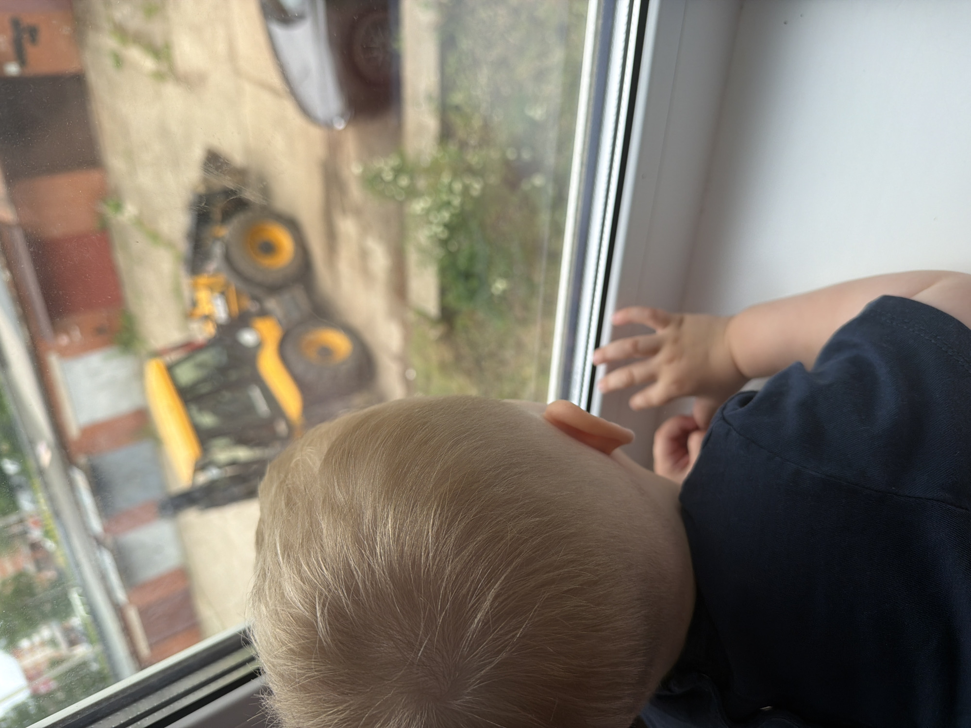 The image displays a fair-haired child, likely a toddler, viewed from an overhead perspective, looking intently out a window. The child's head, with fine blonde hair, and the upper part of a navy blue shirt are visible in the foreground. Their hands rest near the white window frame, suggesting engagement with the view beyond.
Through the window, an outdoor scene unfolds under daylight conditions. A prominent yellow excavator or construction vehicle is visible on what appears to be a paved or dirt area. Due to the camera's angle and the vertical orientation of the exterior view through the window, the excavator appears tilted within the frame. A dark-colored vehicle is also partially visible in the upper section of the outdoor scene. In the background, there are hints of brick-colored buildings or houses, along with green foliage and trees, indicating an urban or suburban environment. The child appears curious and focused on observing the outdoor activity, particularly the construction vehicle. The setting for this observation is Votkinsk, Russia. No visible text is present in the image.