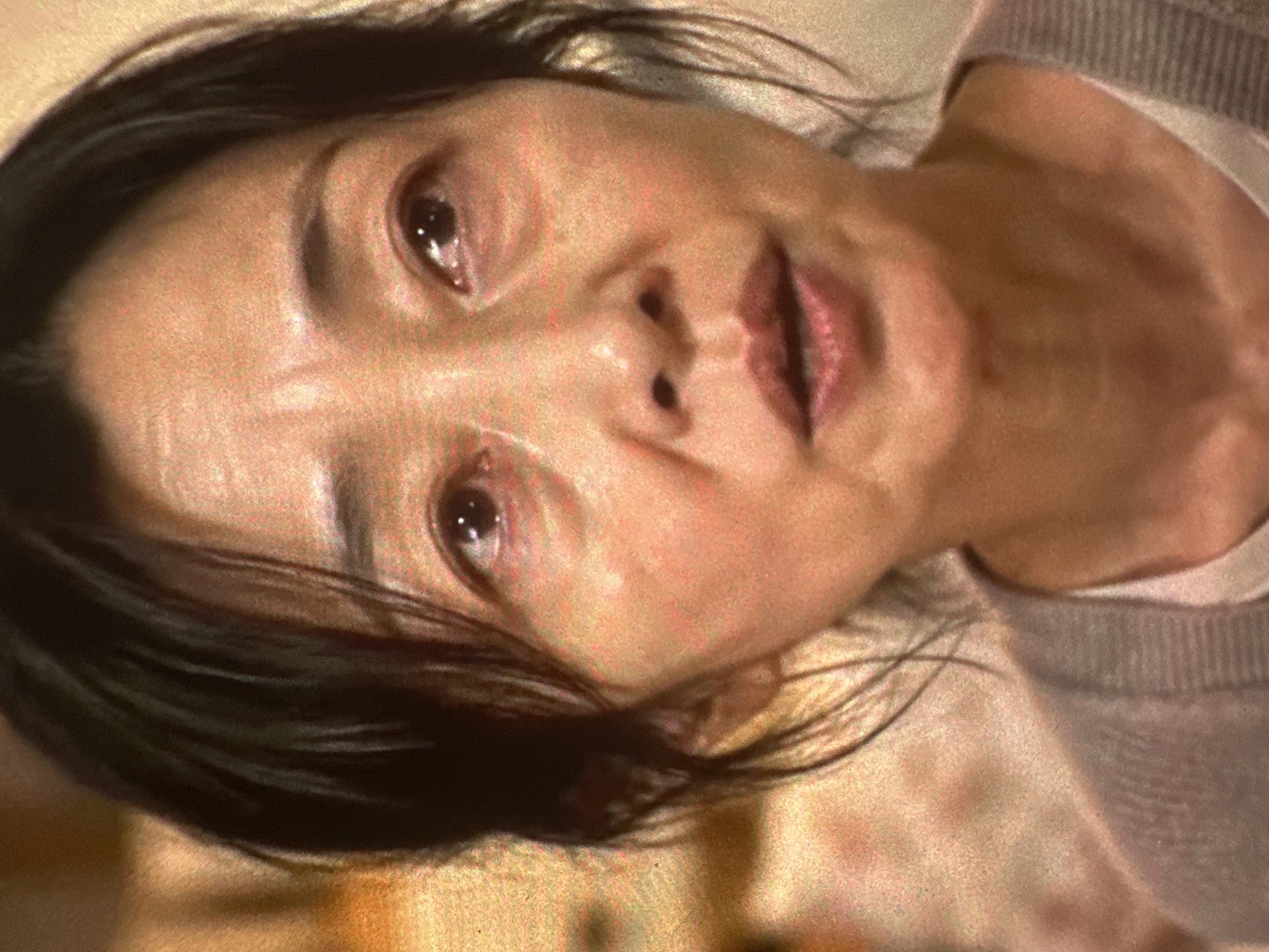 An extreme close-up shot shows the face of an East Asian woman with dark hair, looking slightly to her right with a distressed expression. Her eyes are watery, and her mouth is slightly open as if speaking or about to speak. The lighting is warm and somewhat diffused, casting a soft glow on her skin. The background is out of focus and appears to be an interior setting with textures and colors that suggest a room. The image is grainy, indicating it might be a photograph taken in low light or a still from a video. No specific location in a city is discernible.