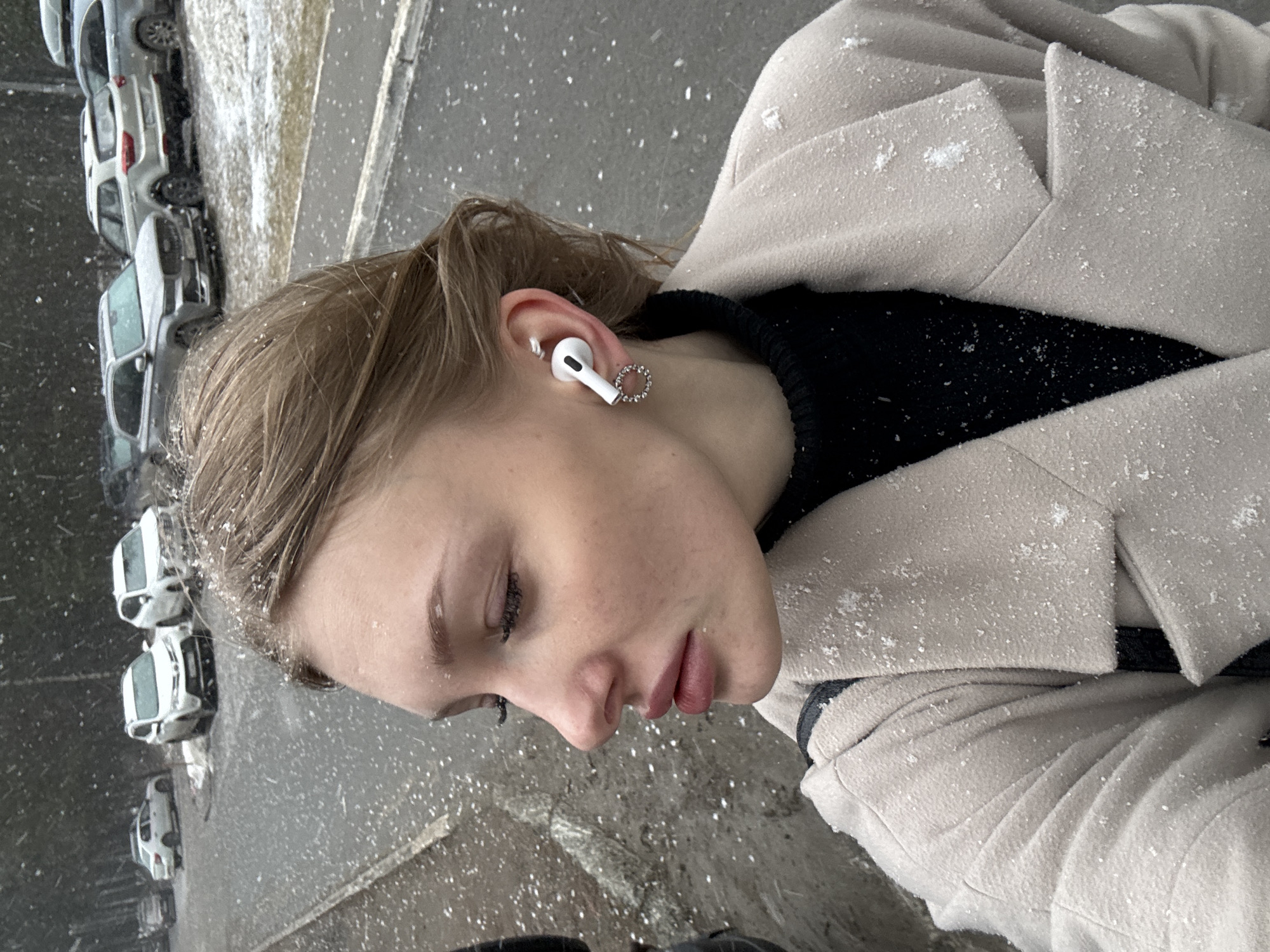 A woman stands outside, presumably in Perm, Russia, given the presence of cars of Russian make. Snowflakes are visible on her clothing, which consists of a light-colored jacket and a black top. She is wearing an earbud in her left ear with a small earring attached. The background shows a city street with parked cars.