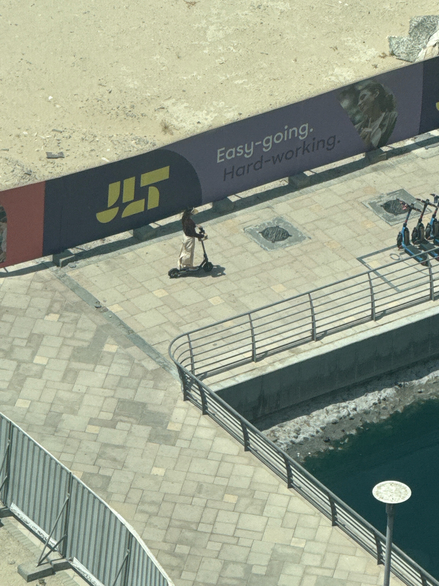 The image shows an outdoor scene in Dubai, United Arab Emirates, likely in the Jumeirah Lakes Towers (JLT) district, indicated by a banner with the JLT logo and text "Easy-going. Hard-working." A person wearing a headscarf and loose-fitting light-colored clothing is riding a black electric scooter on a paved walkway. The walkway is made of light-colored rectangular tiles and is bordered by a metal railing. In the background, there is a body of water, possibly a canal or marina, with several parked electric scooters in a designated area. The environment appears to be a public recreational space with a modern urban setting.