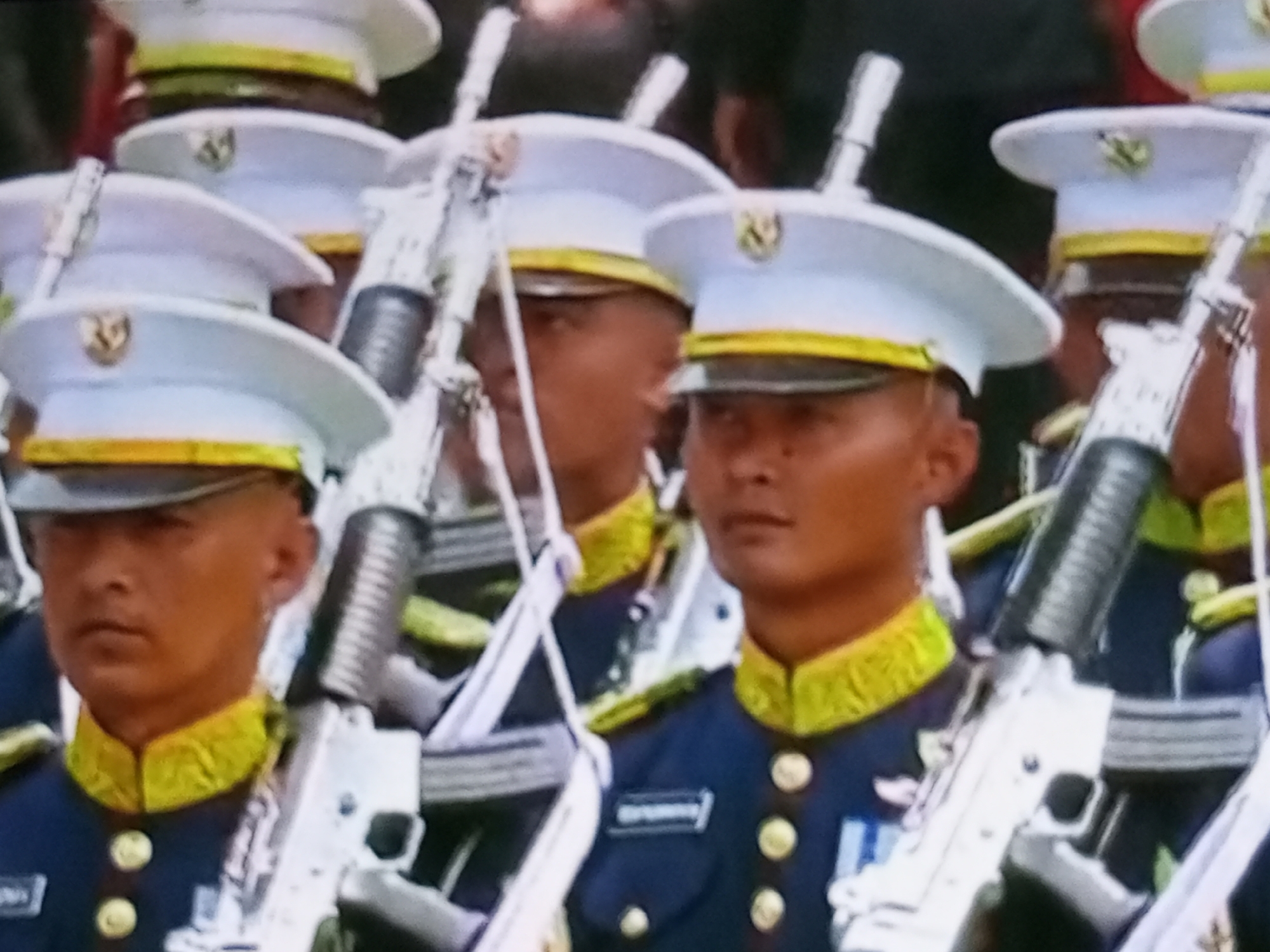 A group of men in ceremonial military uniforms stand at attention holding rifles. The men wear white caps with gold trim and a circular emblem, dark blue jackets with gold trim around the collar and gold buttons, and some wear medals. The rifles are light-colored, possibly silver or white, with black grips and barrels. The men appear to be participating in a parade or ceremony. The background is out of focus, but suggests an outdoor setting with other people present. There is no specific information visible in the image to indicate the location as Banyudono, Indonesia.