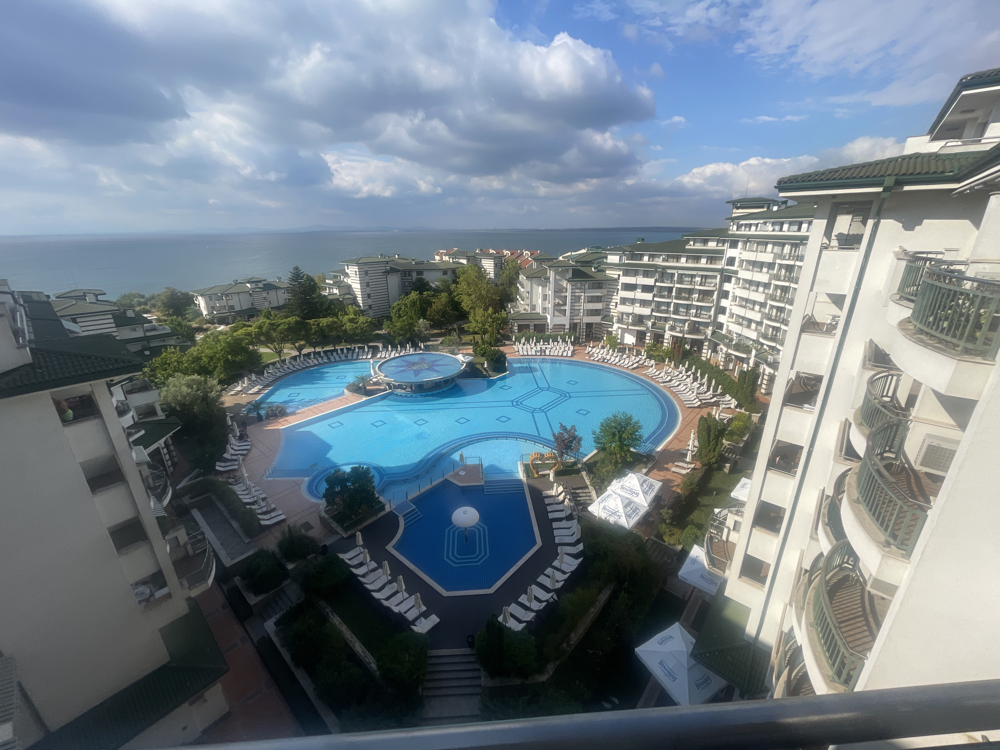 The image shows a resort complex in Ravda, Bulgaria, with a large, ornate swimming pool in the foreground. The pool is surrounded by lounge chairs and umbrellas, and there are several resort buildings visible in the background. The sea is visible in the distance, and the sky is partly cloudy. The resort appears to be well-maintained and offers a relaxing atmosphere.