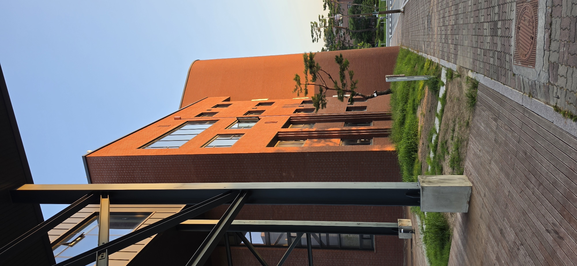 An exterior, low-angle shot depicts a brick building with a curved section on the right, bathed in the warm light of a setting sun. The building, constructed with orange-red bricks, features multiple windows, some of which are open. A thin pine tree stands in front of the curved section of the building. In the foreground, a wooden deck runs parallel to a paved walkway made of interlocking stones. The walkway has a manhole cover and a street lamp visible in the distance, where other buildings and trees can be observed. The sky is clear and blue, suggesting a pleasant evening in Cheongju-si, South Korea.