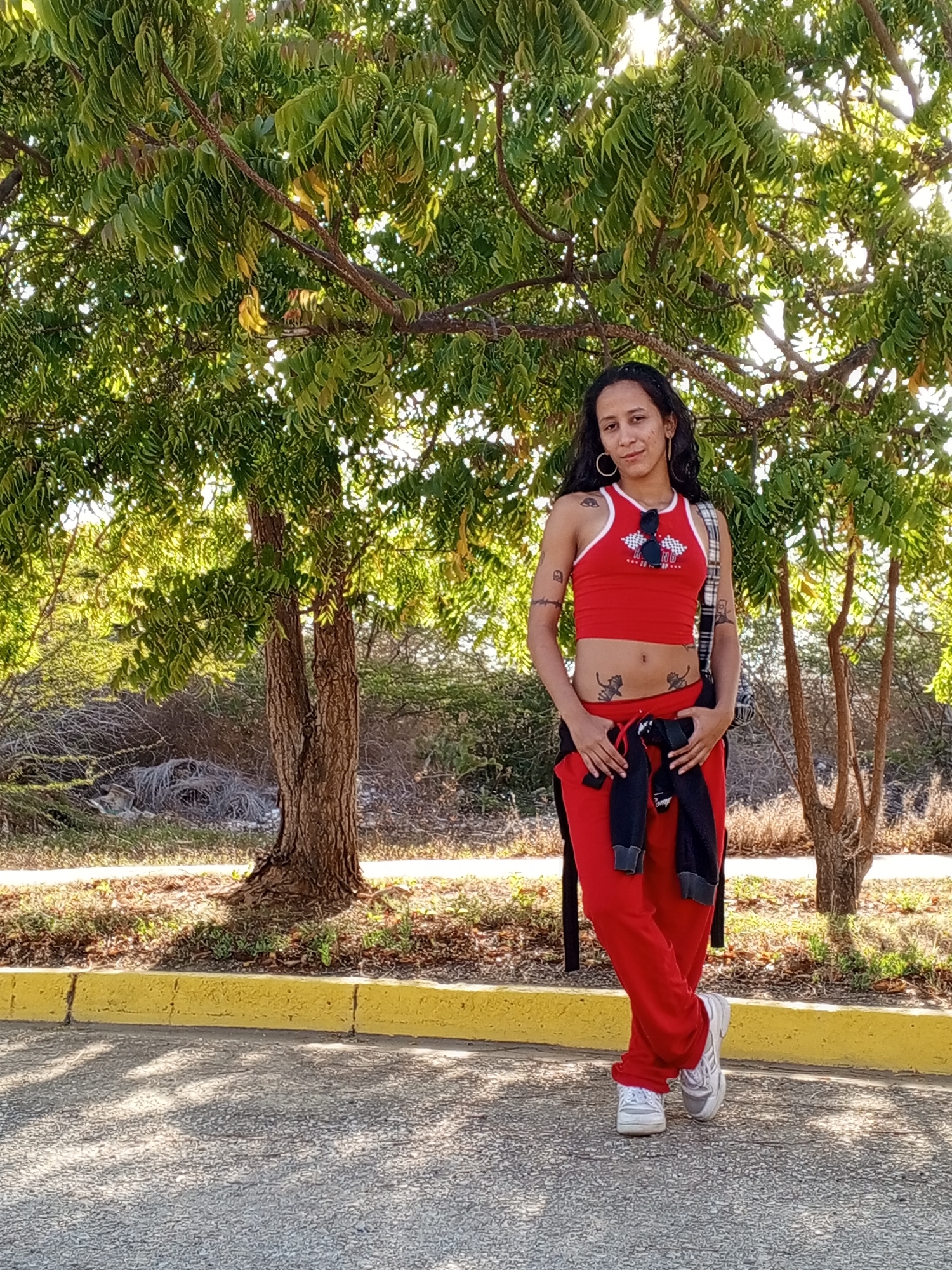 A full-body outdoor portrait features a woman standing under a large tree with abundant green foliage. She has long, dark hair, large hoop earrings, and a neutral to slightly smiling expression. Her attire consists of a vibrant red athletic-style outfit: a sleeveless crop top with white trim and a graphic print featuring a checkered flag and the letters "RD," paired with matching red track pants. A black garment, possibly a sweatshirt or jacket, is tied around her waist, and she wears white and gray sneakers. Sunglasses are hooked onto the neckline of her top.
The woman displays several tattoos, including a geometric or tribal design on her upper left arm, band-like markings on her right arm, and three small dark figures (possibly cats) on her lower abdomen.
The setting suggests a sunny day, with bright light filtering through the tree canopy and casting shadows on the asphalt ground. In the foreground, a distinct yellow painted curb separates the paved surface from the base of the tree and some sparse ground cover. The background consists of additional trees and dry, scrubby vegetation, indicating a warm, possibly arid, outdoor environment. The woman is posing for the camera, projecting a confident demeanor.