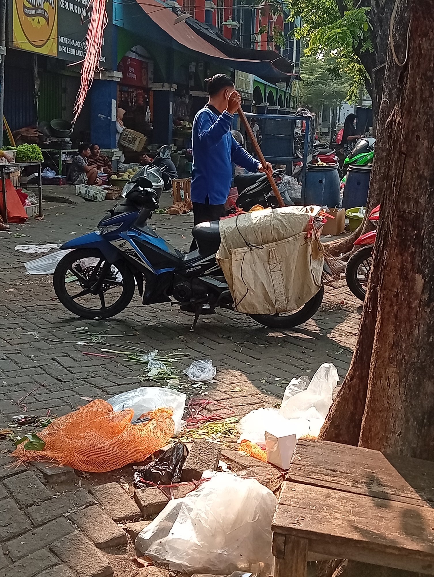 This is a vertical, medium shot photograph taken at a street market in Larangan, Indonesia. The scene depicts a man wearing a blue long-sleeved shirt and dark pants, holding a broom and standing next to a parked blue motorcycle. The motorcycle has a large, tan bag strapped to the rear. In the foreground, scattered on the brick pavement, are various discarded items including an orange mesh bag, white plastic bags, and some plant debris. Behind the man and the motorcycle, a busy market atmosphere is evident with stalls, other motorcycles, and several people. The lighting suggests it is daytime, with shadows cast on the ground. The overall impression is that of an outdoor marketplace with activity and discarded waste.