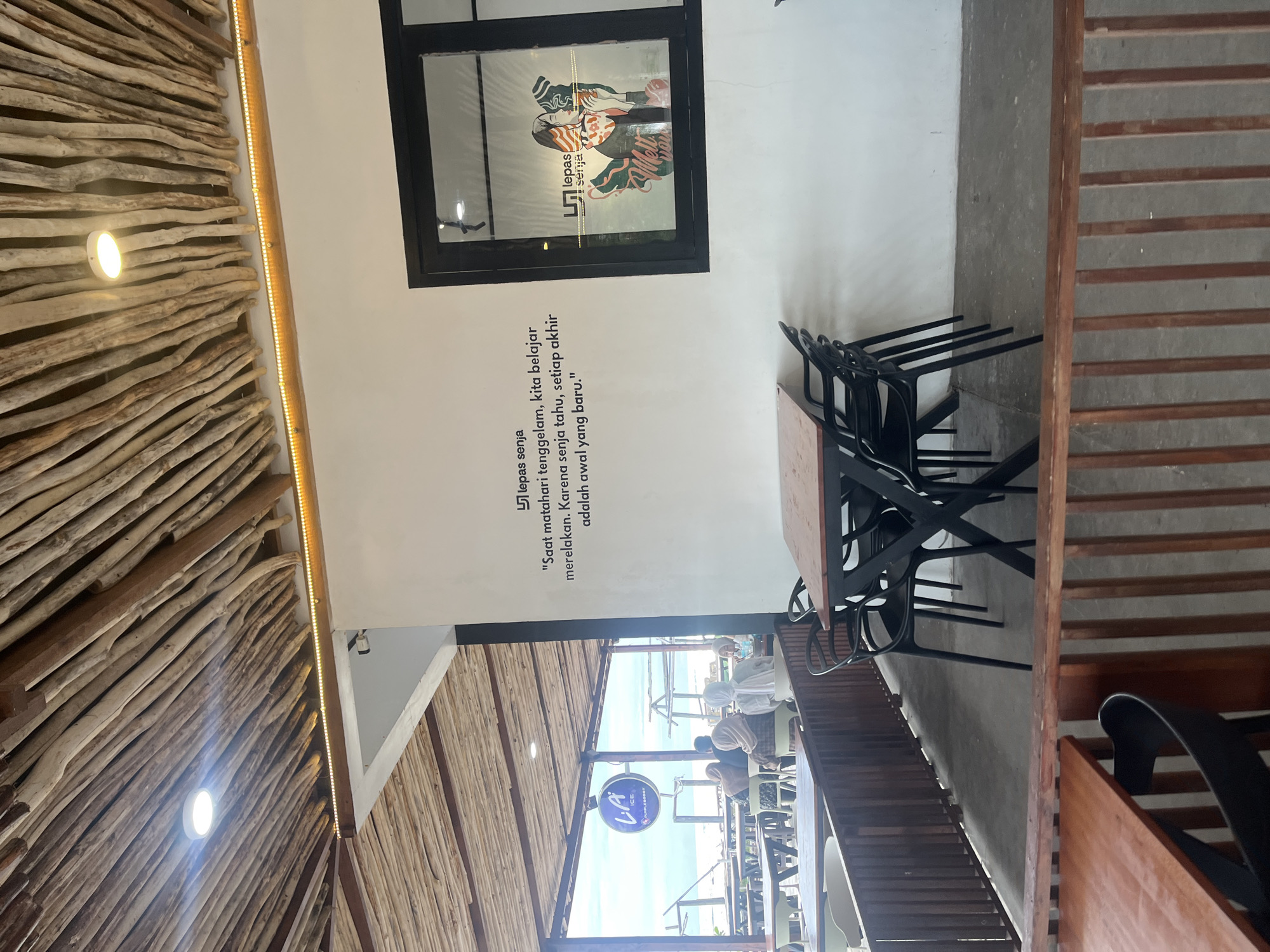 The image depicts an outdoor seating area of a cafe, likely located in Monikeun, Indonesia, with a rustic, natural aesthetic. The ceiling is constructed from a lattice of natural wood branches, with a round white light fixture and a strip of warm-toned LED lights running along the edge of the white wall.
On the white wall, there is a quote in Indonesian: "Saat matahari tenggelam, kita belajar merelakan. Karena senja tahu, setiap akhir adalah awal yang baru." (When the sun sets, we learn to let go. Because dusk knows, every end is a new beginning.) To the right of the quote, a window looks into the interior of the cafe, where a graphic featuring two figures holding drinks is visible, along with the cafe's logo, "lepas senja."
In the foreground, there are several dark-colored tables and chairs arranged on a concrete floor. A wooden railing separates the seating area from a lower level, where more tables and chairs are partially visible. In the background, beyond the railing and tables, a view of the ocean and sky can be seen, suggesting a coastal location. Several patrons are seated at tables in the background, facing the ocean. One prominent circular sign with the text "LA ICE" and "PURPLE BOOST" is visible in the distance, possibly indicating the cafe's name or a featured product. The overall atmosphere appears relaxed and casual, possibly a beachside cafe.