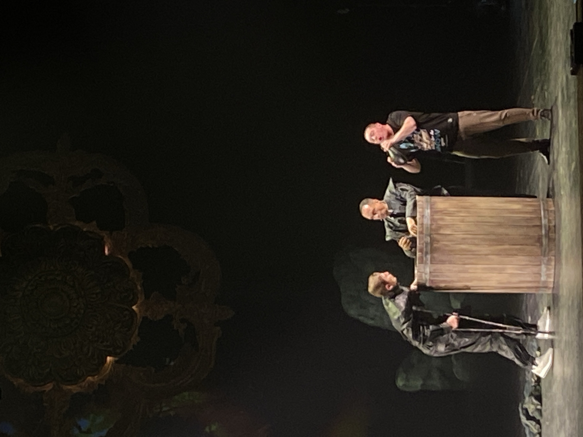 A dimly lit stage, likely a theater, in Unknown, Unknown, shows three men engaged in a performance. On the left, an ornate decorative element is visible. The bottom man, dressed in a dark suit and white shoes, stands holding crutches. He faces a man behind a wooden barrel. A third man, in a t-shirt and khaki pants, stands above. The scene is obscured by darkness.