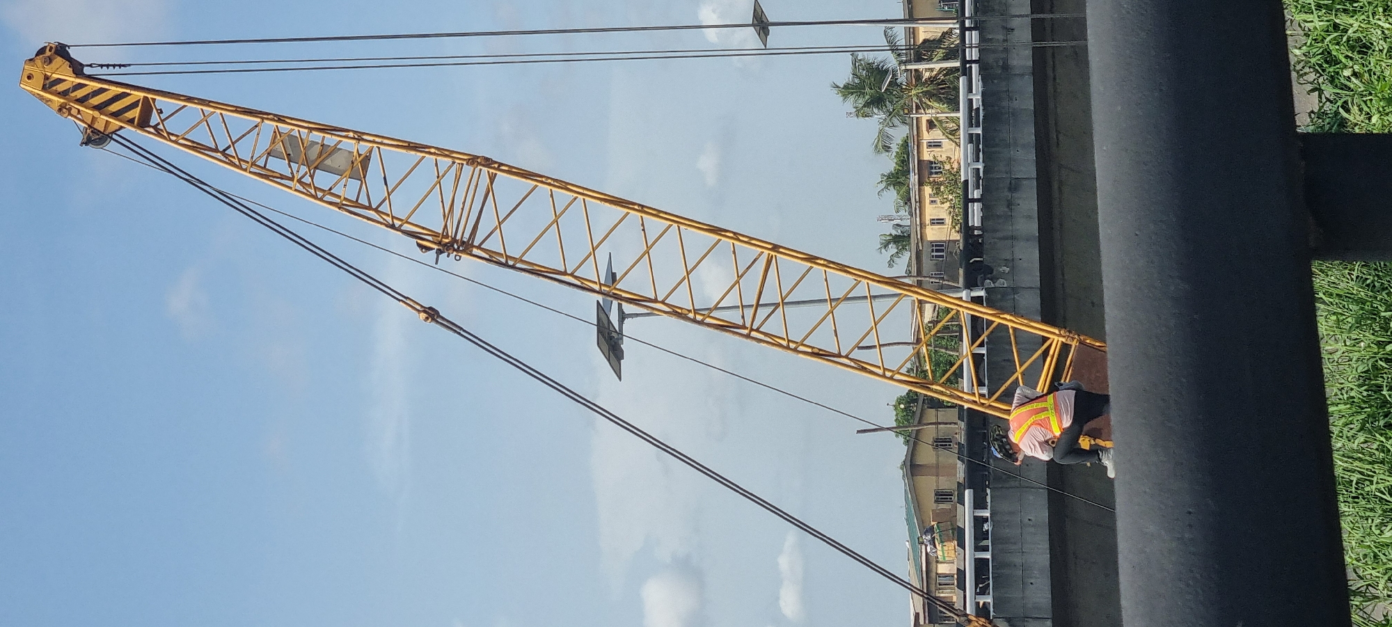 The image shows a yellow lattice boom crane against a partly cloudy sky, likely in an urban area. The crane is angled upward and leftward. A worker wearing a safety vest and helmet is visible near the base of the boom. Buildings with palm trees are visible behind the crane. A black cylindrical structure (possibly a pipe or pillar) is in the foreground, partially obscuring the view. Based on the location information, this scene is likely in the city of Satellite Town, Nigeria.