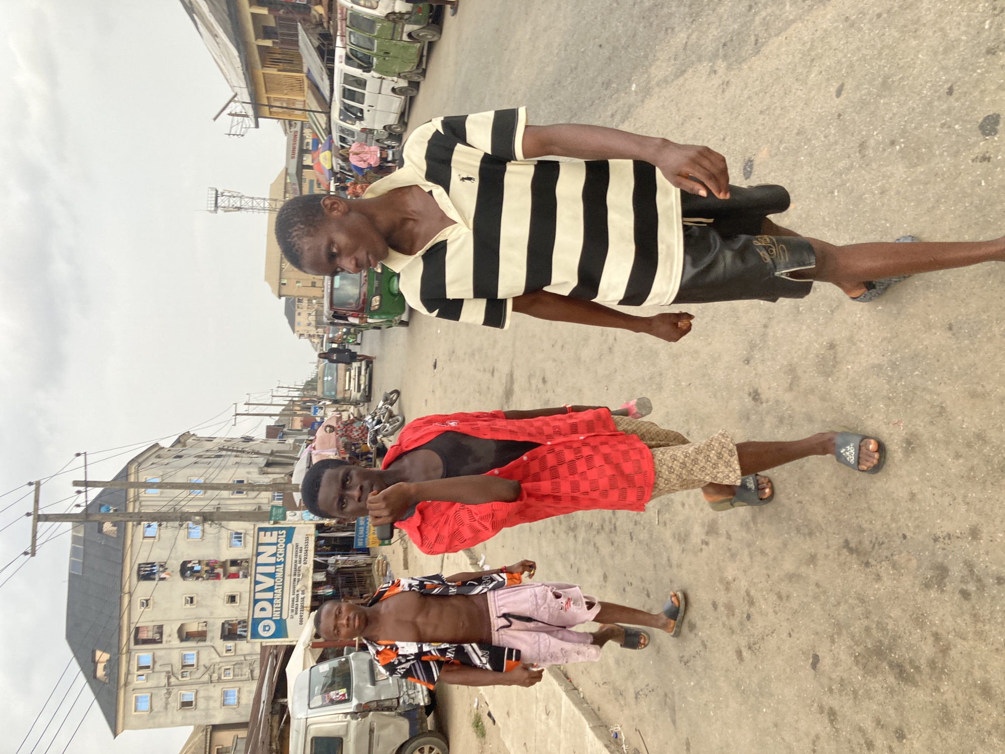 The image shows three young men on a street in what appears to be Ura Nta Umuarandu, Nigeria. The man at the top is wearing a black and white striped polo shirt and dark shorts. The middle man wears a red shirt and patterned shorts. The bottom man is bare-chested with a patterned shirt and pink shorts. They are all wearing sandals. Behind them, there is a sign for Divine International Schools on a building. The street scene includes various vehicles and street infrastructure.