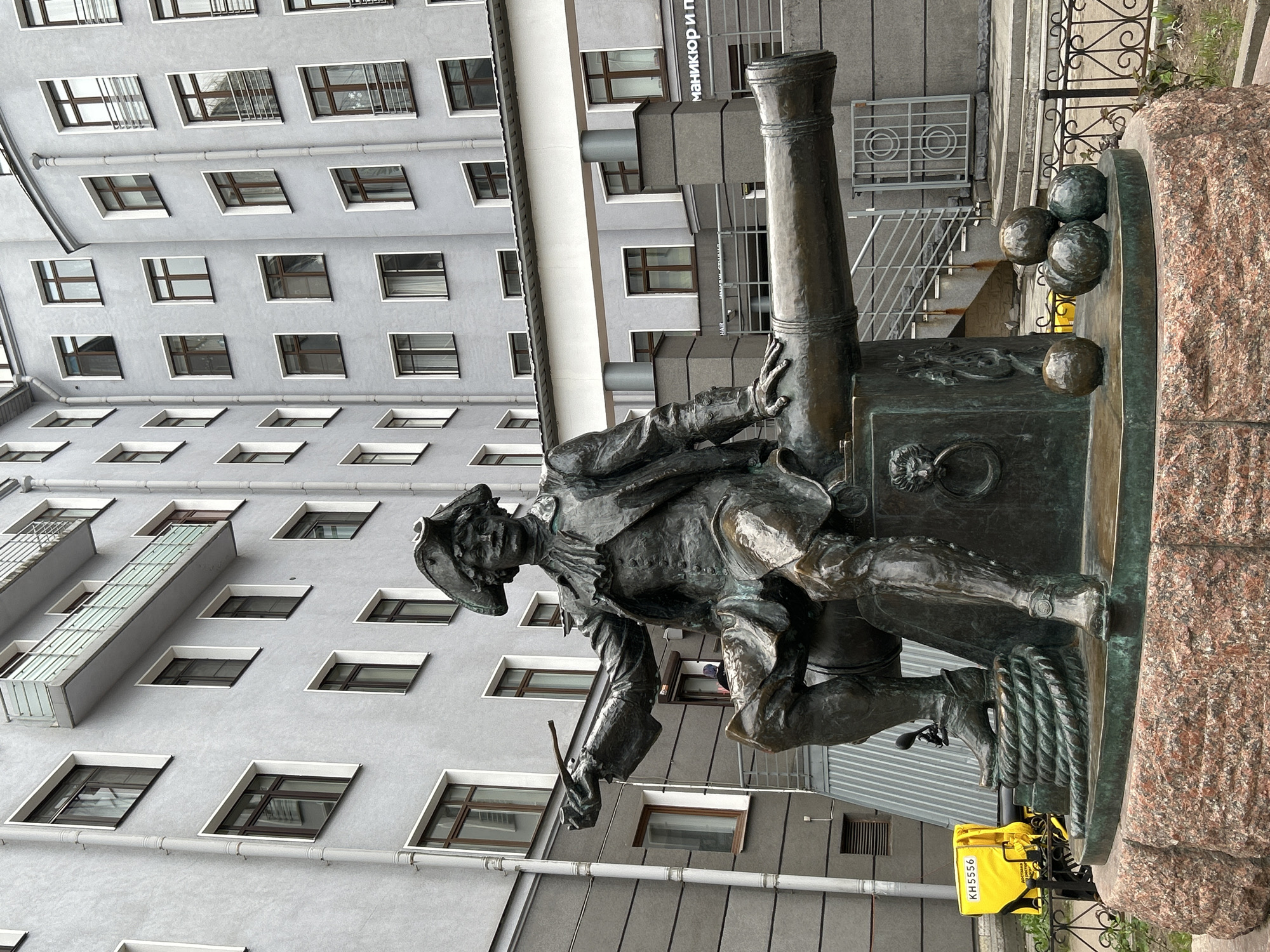 A statue of a man sitting next to a cannon is in Saint Petersburg, Russia. The statue depicts a man in historical attire, seated next to a cannon with cannonballs. The statue is bronze-colored. Behind the statue is a multi-story building with many windows. There is also a sign written in Cyrillic and a staircase with a metal railing. A yellow delivery bag is visible in the lower right of the frame.