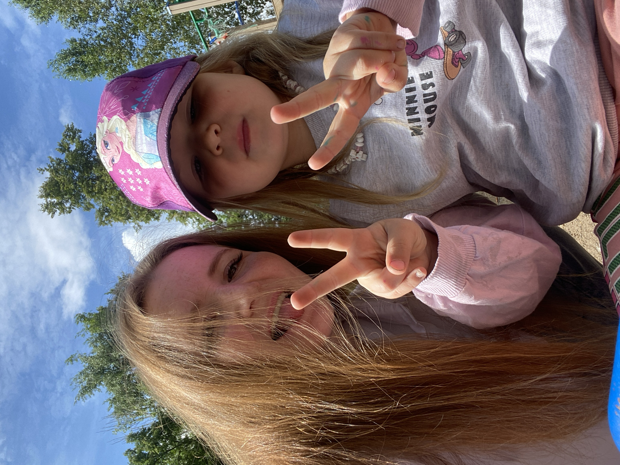 Two females, one older with long, reddish-brown hair and the other a young child wearing a pink cap with an image of Elsa from Frozen, are posing for a selfie outdoors. Both are making a peace sign with their fingers. The older female has her tongue sticking out. The child is wearing a grey sweatshirt with a Minnie Mouse graphic. They appear to be in a park or playground setting, with trees and a blue sky with clouds in the background. The setting is consistent with outdoor recreational areas found in cities like Chaikovsky, Russia, which has numerous parks and green spaces.