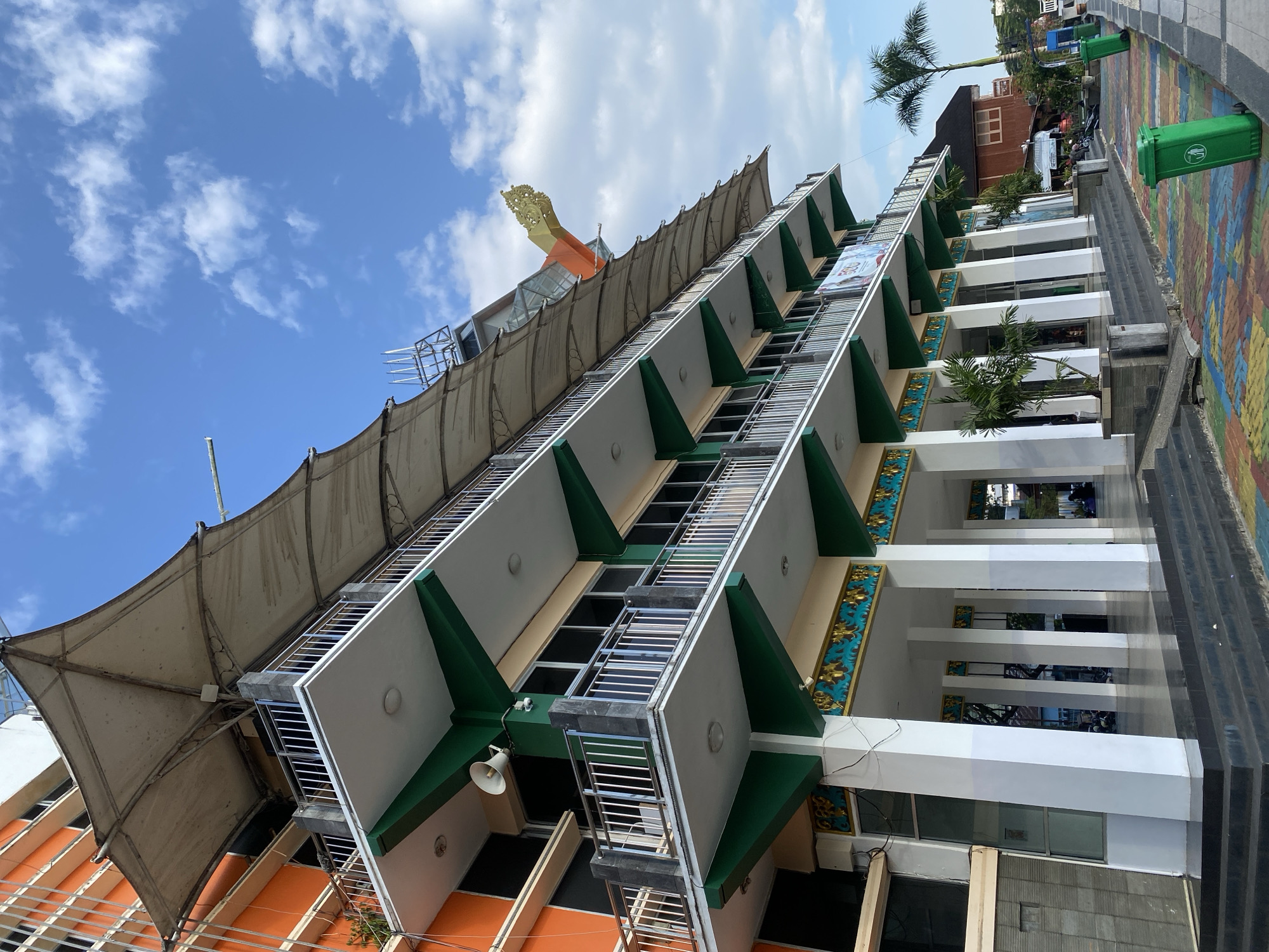 The image depicts the exterior of a multi-story building with a distinctive cantilevered awning system in Banjarmasin, Indonesia. The building has an orange facade on the left and layered balconies with white railings and green trim. The awning is a light-colored, fabric structure supported by metal poles, creating a wavy effect over the balconies. Some decorative elements, including golden floral motifs, are visible below the balconies. The ground level features white pillars and a walkway with colorful paving stones. Two green trash cans are placed along the walkway. In the background, a palm tree and other buildings are visible. The sky is blue with scattered white clouds. There are no people clearly visible in the foreground or mid-ground, though some figures may be present further in the background.