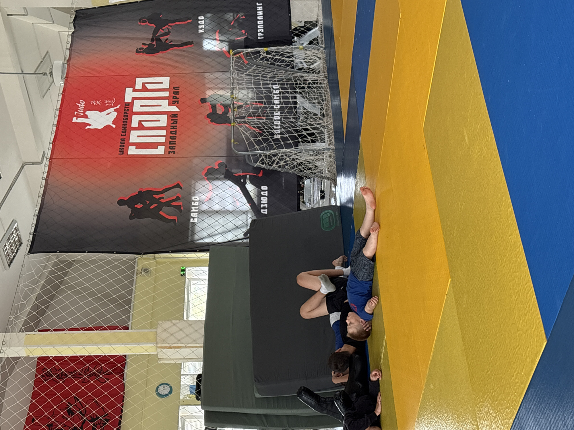 This image captures an indoor martial arts training facility, likely in Chaikovsky, Russia, given the location context and visible text. The scene is dominated by a large area of blue and yellow matting, arranged in a tatami-style pattern, where several children are engaged in training.
In the foreground, two children are on the blue and yellow mat. One child, wearing a blue top and patterned shorts, is on their back, while another child, in dark clothing, appears to be grappling or practicing a hold on top of them. Another child is partially visible on the far left edge, also in dark attire. Further back, on a section of dark grey crash mats, another child is lying down. The children are barefoot, typical for martial arts practice.
The background features a large red and black banner covering a significant portion of the wall. This banner displays silhouettes of martial artists performing various techniques. Prominently featured in white text is "СПАРТА" (SPARTA), with "ШКОЛА ЕДИНОБОРСТВ" (School of Martial Arts) above it and "ЗАПАДНЫЙ УРАЛ" (Western Urals) below. Other visible Russian text next to martial arts silhouettes includes "Judo" (in English script), "КУДО" (Kudo), "ГРЭППЛИНГ" (Grappling), "БОЕВОЕ САМБО" (Combat Sambo), "ДЗЮДО" (Judo), and "САМБО" (Sambo), indicating a range of combat sports taught at the facility. White safety netting encloses part of the training area. A window, a wall clock, and general indoor lighting are also visible, suggesting a daytime training session.