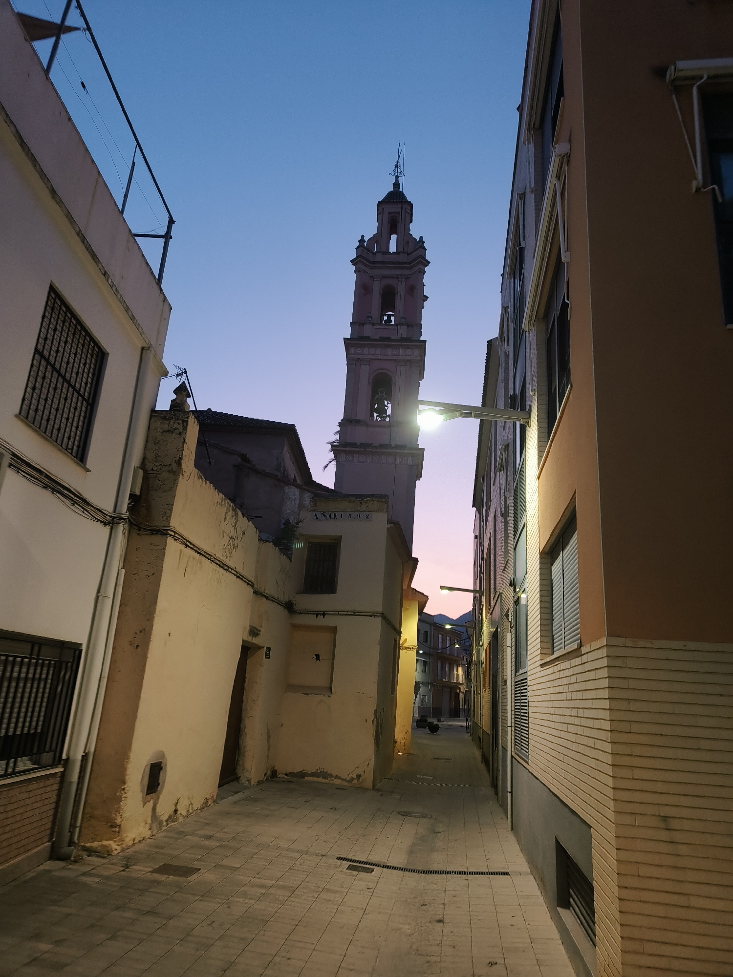 The image depicts a narrow street in Benirredrà, Spain, at dusk. The sky is a gradient of deep blue at the top transitioning to pale pink and orange near the horizon. Dominating the scene is a tall, ornate bell tower, likely part of a church, with architectural detailing and a bell visible in one of its openings. The tower is made of a light-colored stone, possibly pinkish hue, and is illuminated by artificial light.
Flanking the street are buildings on either side. The building on the left is lighter in color with windows featuring dark metal grates. The building on the right is a warm peach color with brick detailing on the lower portion. This building has multiple windows, some with horizontal blinds, and external light fixtures are mounted on its facade.
The street itself is paved with light-colored square tiles and features drainage grates running along its sides. The perspective suggests the camera is looking down the street, away from the viewer, towards the distant buildings and the colorful sky. There are no people visible in the image, and the scene appears quiet and serene. The overall impression is one of a charming European street bathed in the soft light of twilight. The date "ANO 1892" is visible on the side of a building closer to the tower.