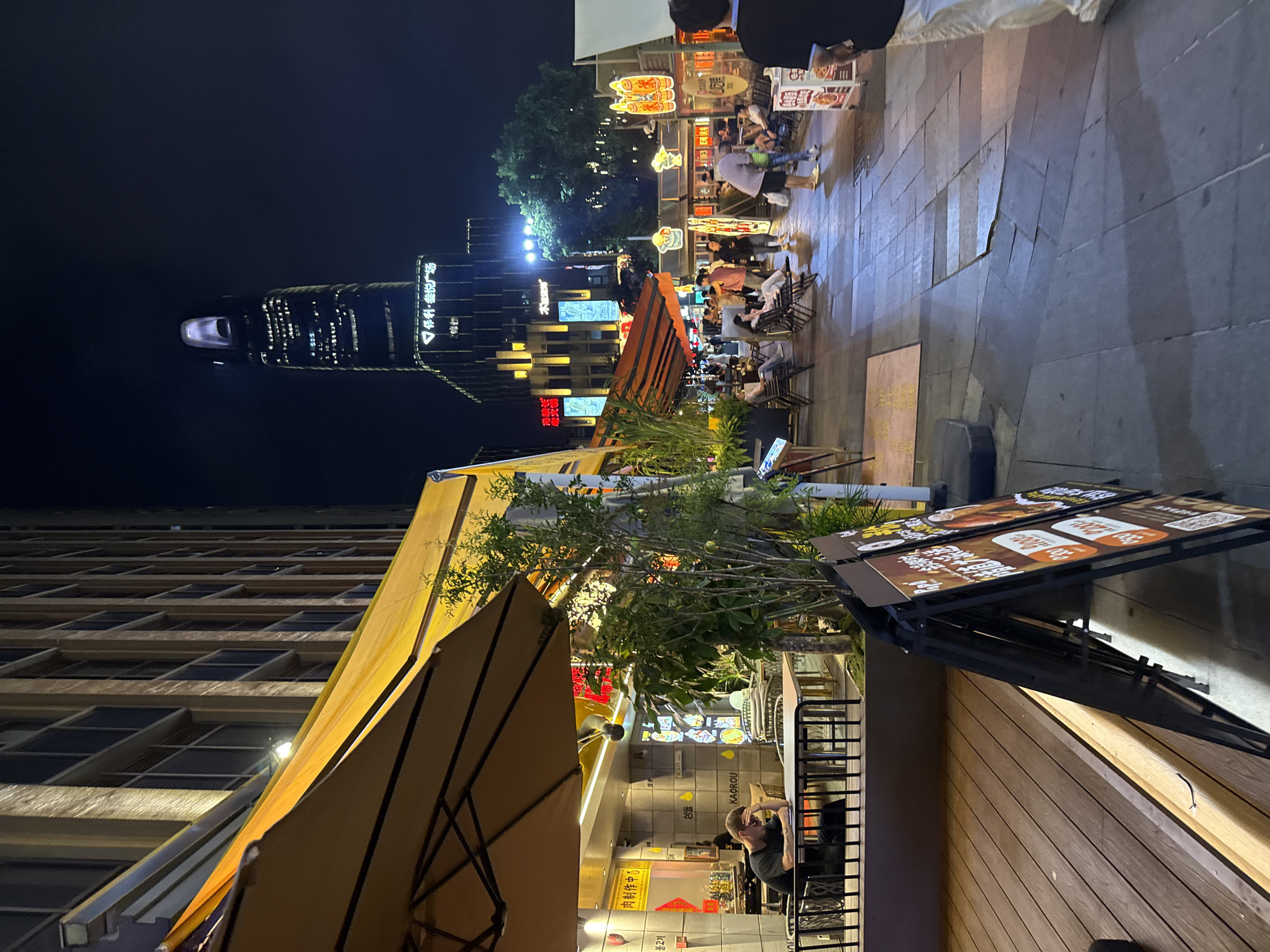 The image shows an outdoor scene in Haizhu District, China, at night. There are several restaurants with outdoor seating areas. Some patrons are seated at tables, and others are walking by. Signage and awnings are illuminated by artificial lights. A tall skyscraper is visible in the background. A man sits at an outdoor table, partially obscured by a large umbrella. A food menu is displayed on a stand near the restaurant. There is a building with a grid-like facade to the side of the restaurant. The pavement is made of large, rectangular stones.