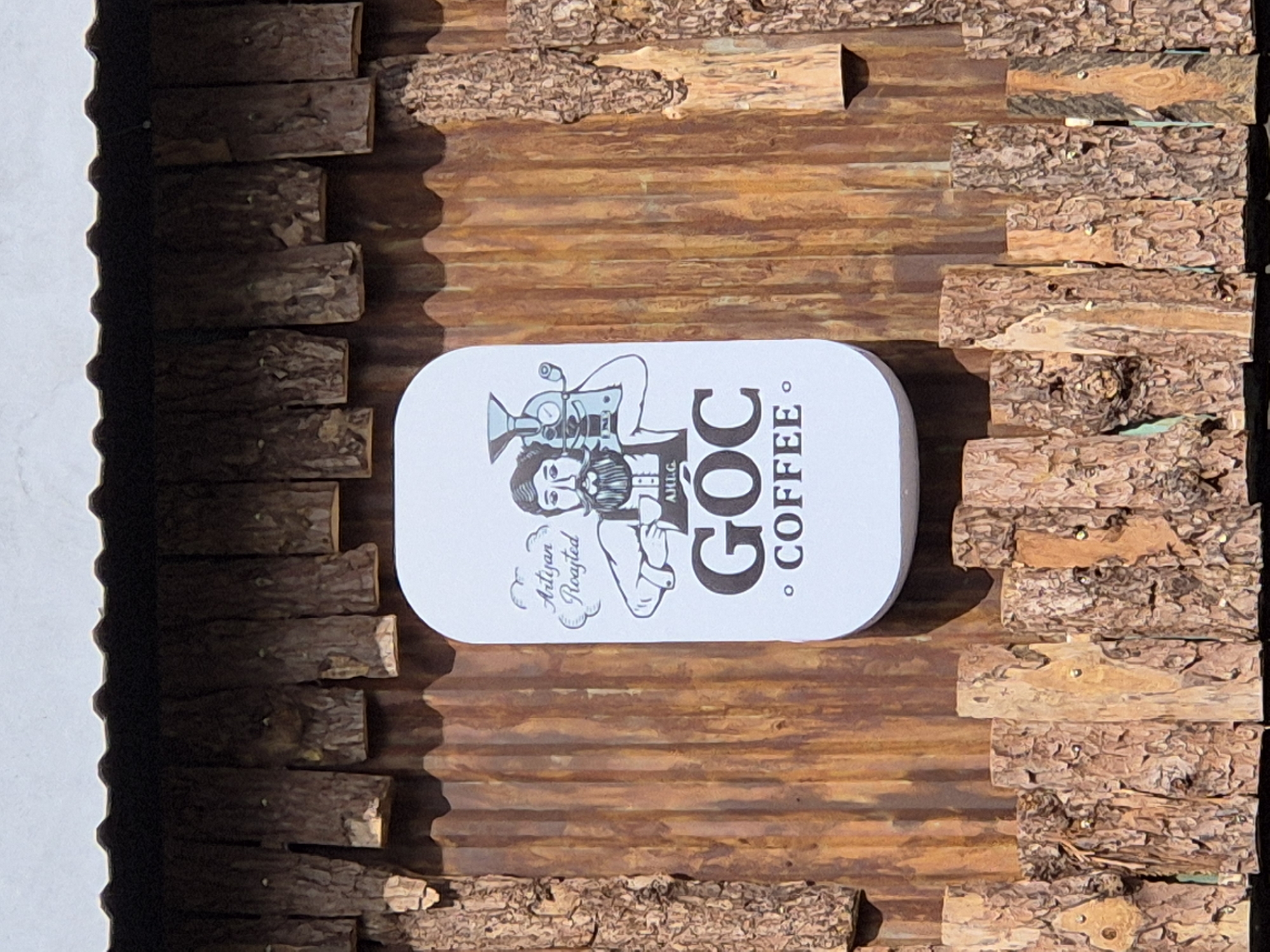A white, oval-shaped sign with a cartoon illustration is affixed to a rustic wooden wall. The sign depicts a man with a beard wearing an apron, holding a coffee roaster. Above the man, stylized text reads "Artisan Roasted." Below the man, smaller text says "AHL.G." The largest text on the sign reads "GOC COFFEE." The wall is constructed of vertically oriented wooden planks with a corrugated metal texture visible behind the planks. The top and bottom of the wall are adorned with a decorative border of rough-hewn logs. This sign is located outside GOC COFFEE in Da Nang, Vietnam.
