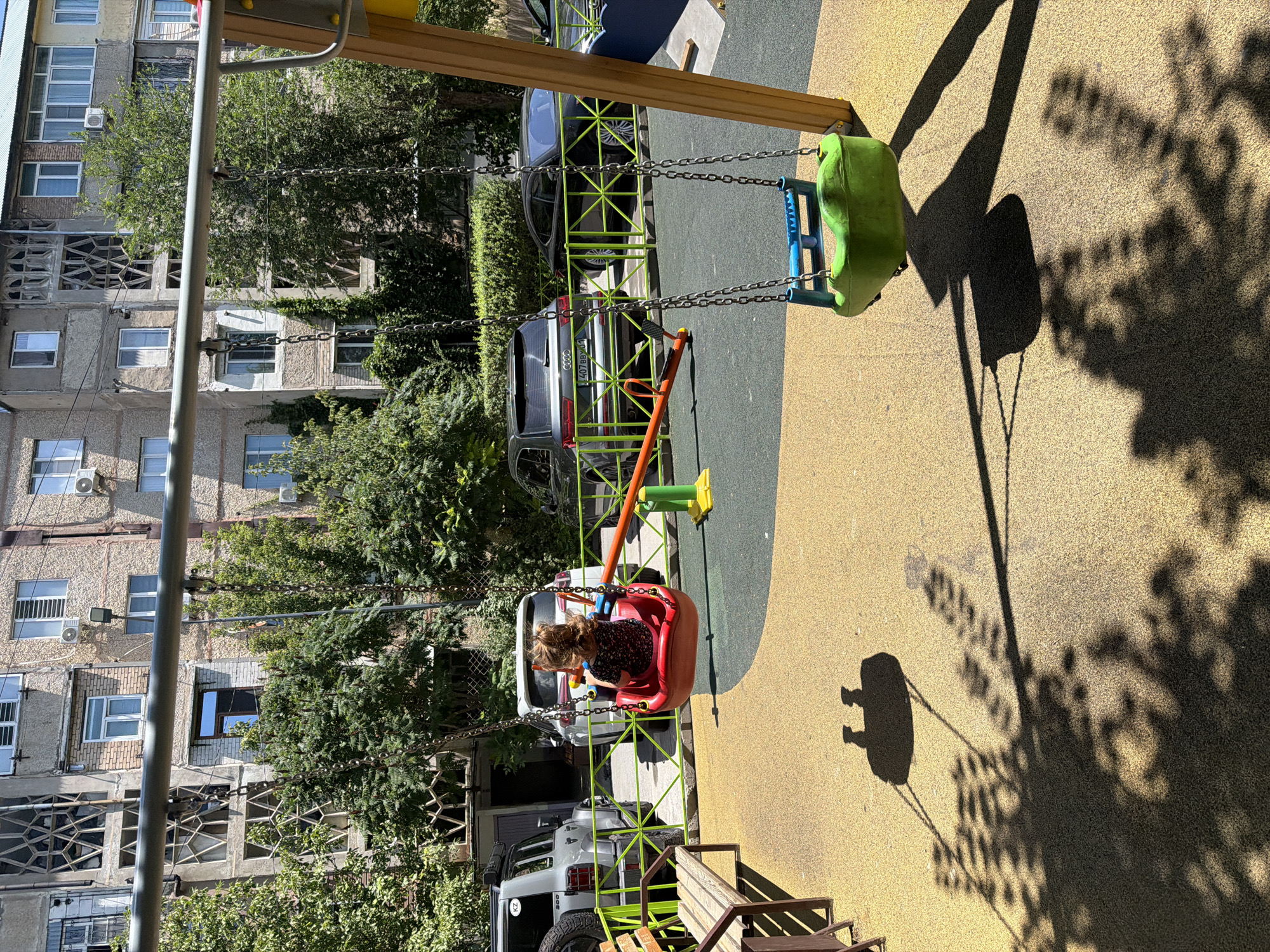 The image depicts a children's playground located in the city of Shymkent, Kazakhstan, adjacent to a residential building. In the foreground, a child sits on a red swing, facing away from the camera. To the right, a green swing hangs from a yellow play structure. A seesaw, with an orange seat and yellow supports, is positioned between the swings. In the background, several cars are parked behind a green fence, with trees and a multi-story apartment building visible beyond them. The playground surface is a textured yellow material.