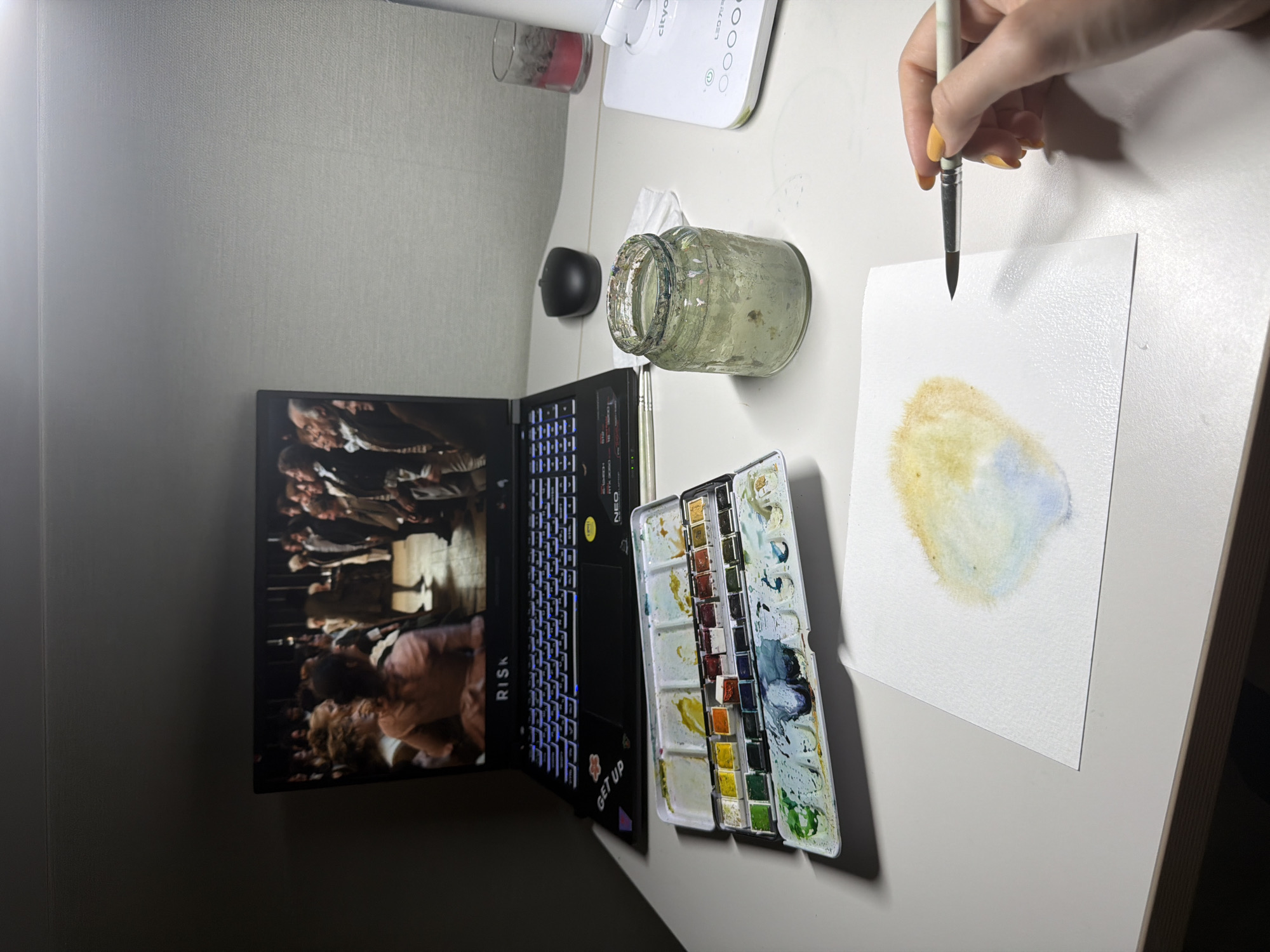 A high-angle medium shot captures a workspace on a desk in Dongducheon-si, South Korea. The desk is set up for watercolor painting, with a laptop open in the background displaying a historical scene. To the left of the laptop is a watercolor palette filled with various colors. In front of the palette, a sheet of watercolor paper shows a splotch of yellow and blue paint. A glass jar filled with murky water sits to the right of the paper, and a mouse is positioned between the laptop and the jar. A desk lamp is visible on the far right, with a red candle holder next to it. A hand with yellow nail polish is holding a paintbrush, poised to add more color to the paper. The overall environment suggests a creative and relaxed atmosphere.