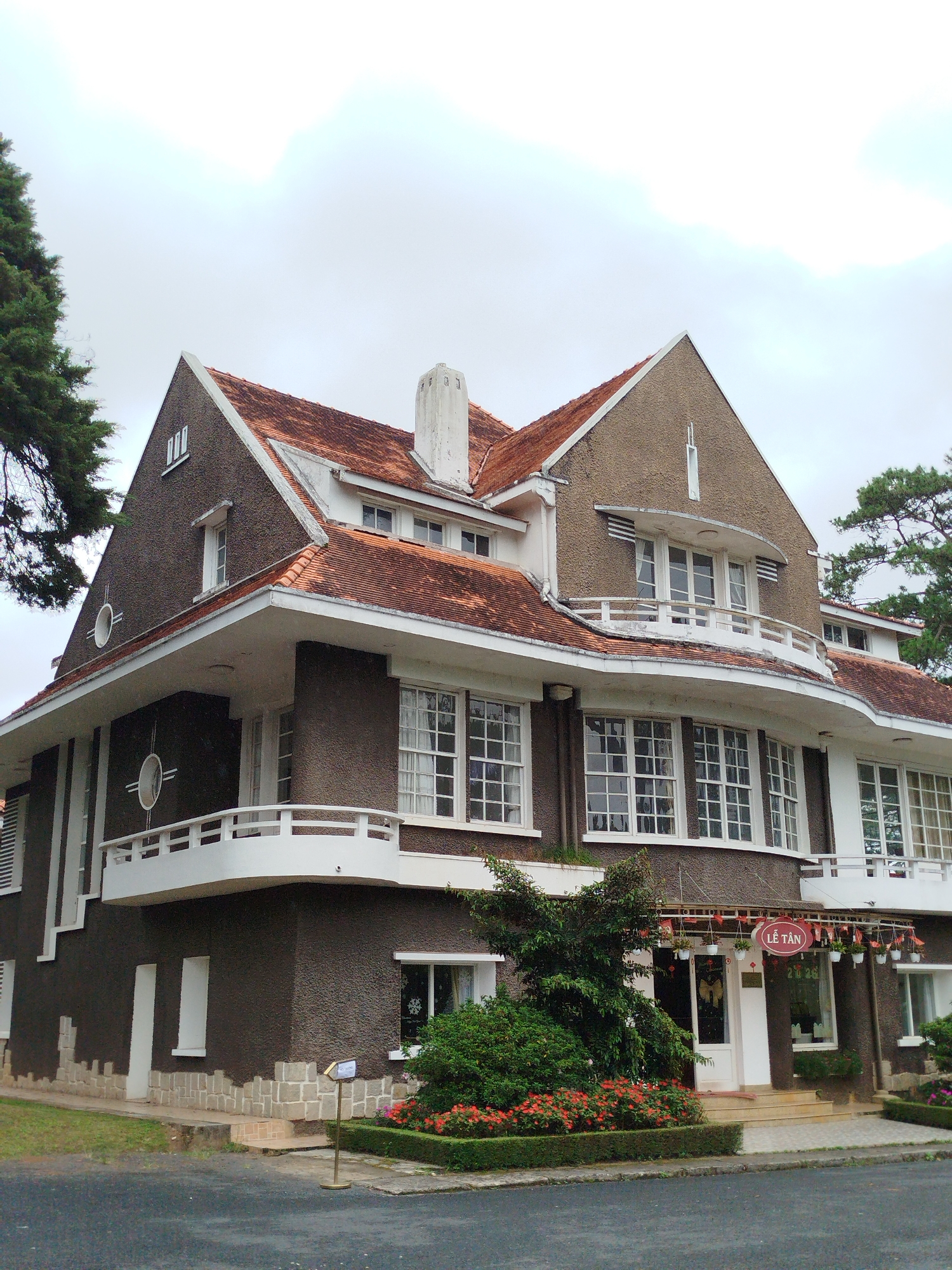 The image shows a large, two-story building with a distinctive architectural style. The building has a dark, textured facade, a red tiled roof, and white trim around the windows and balconies. There are numerous windows, many of which are multi-paned. A prominent white balcony wraps around the curved front of the building on the upper level, and another on the lower level.
In front of the building, there is a small garden area with flowering plants and bushes. A narrow pathway leads to the entrance, which is a white door with glass panels. Above the entrance, a red oval sign with Vietnamese text that appears to say "LÊ TÂN" is displayed, along with hanging decorations and potted plants.
To the left of the building, a large evergreen tree partially obscures the upper corner. To the right, another evergreen tree is visible. The ground in front of the building is paved, and to the left, a grassy area is present. A metal signpost stands in the grassy area. The sky is overcast and light.
This building is identified as the Dinh 2 (Viceroy's Palace) in Da Lat, Vietnam. It's a historic structure known for its French colonial architecture.