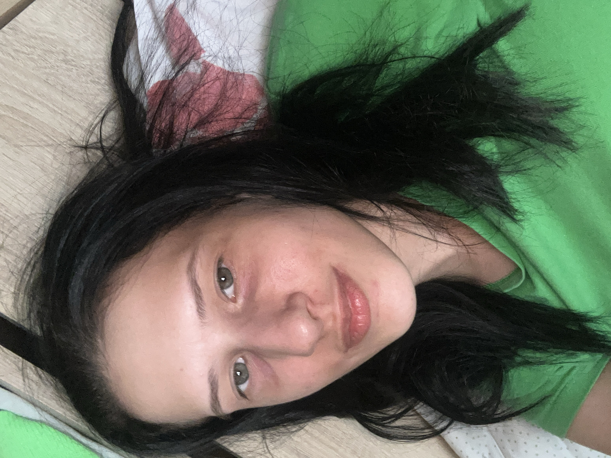 A close-up, overhead shot shows a young woman with dark hair lying on her back, her head tilted slightly to the right. She is wearing a bright green t-shirt. Her eyes are open and looking upwards, and her lips are slightly parted. The background appears to be a wooden surface with a patterned fabric beneath her head. The lighting is soft, creating a natural glow on her skin. The overall impression is one of relaxed repose, possibly taken in a bedroom or living area. The image does not contain any elements that indicate its location in Perushtitsa, Bulgaria.